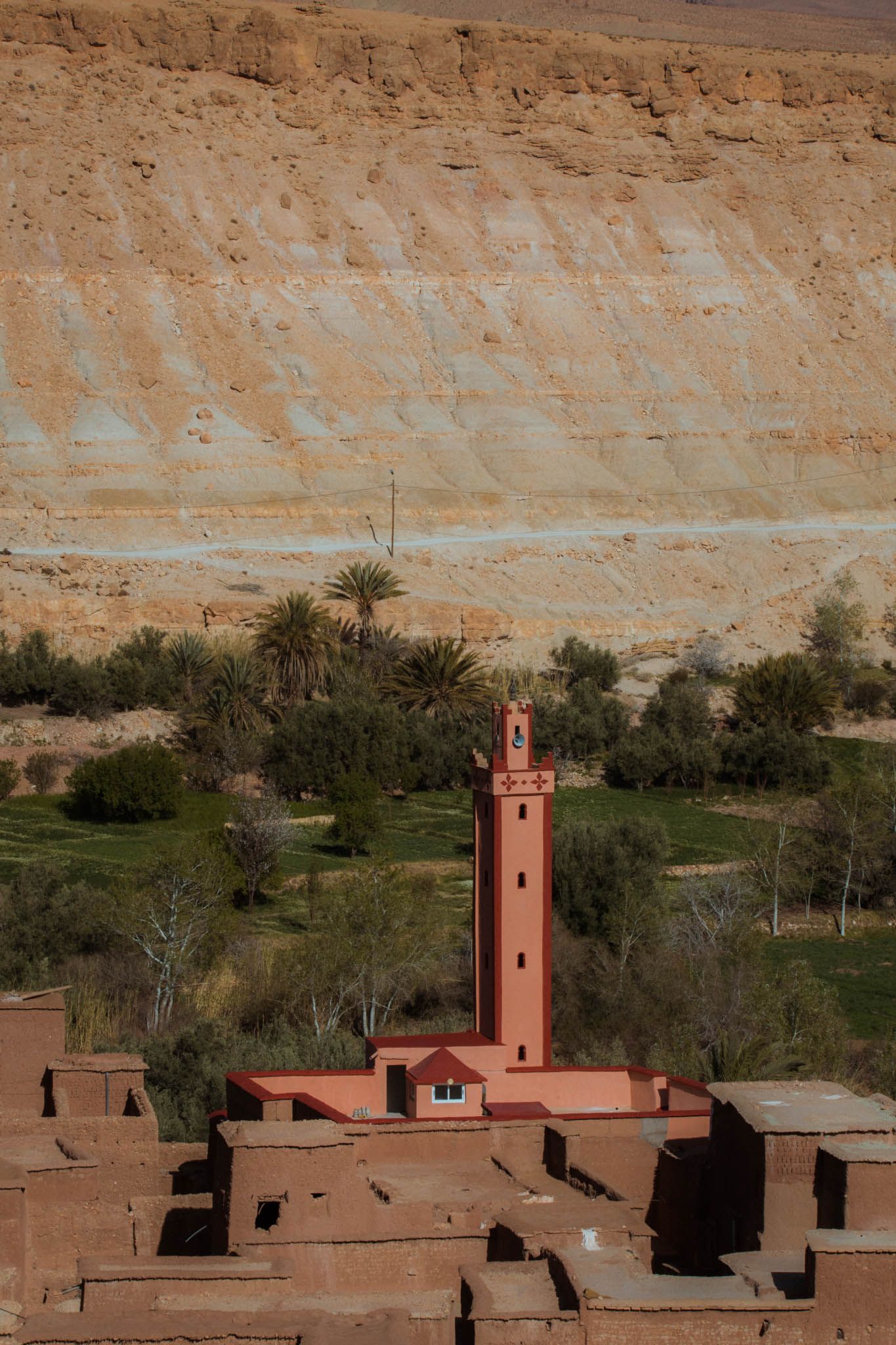 A traditional red clay mosque with a minaret tower stands against desert cliffs in an oasis landscape.