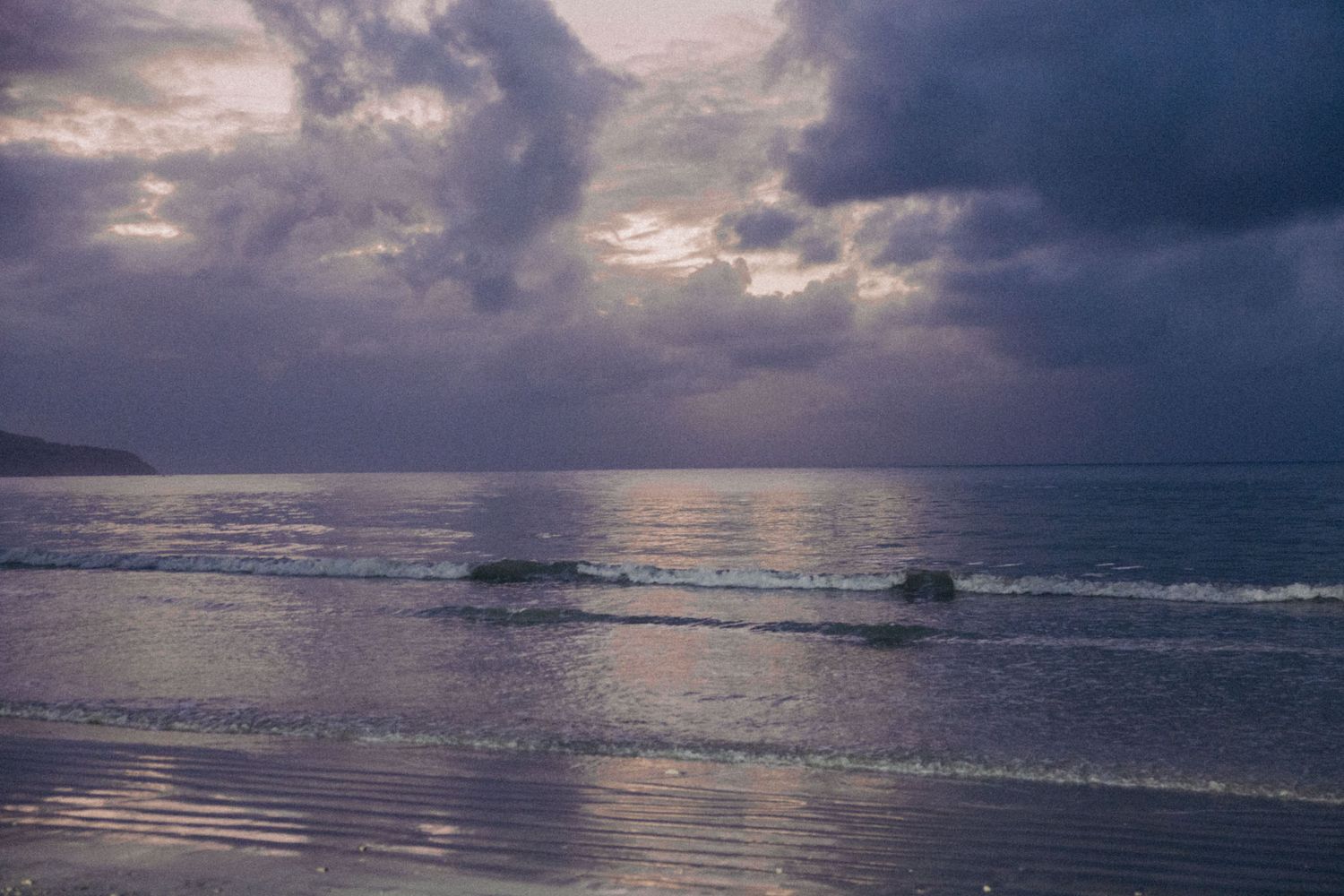 Dramatic cloudy sunset over a serene beach with waves rolling onto shore and sun rays piercing through dark clouds.