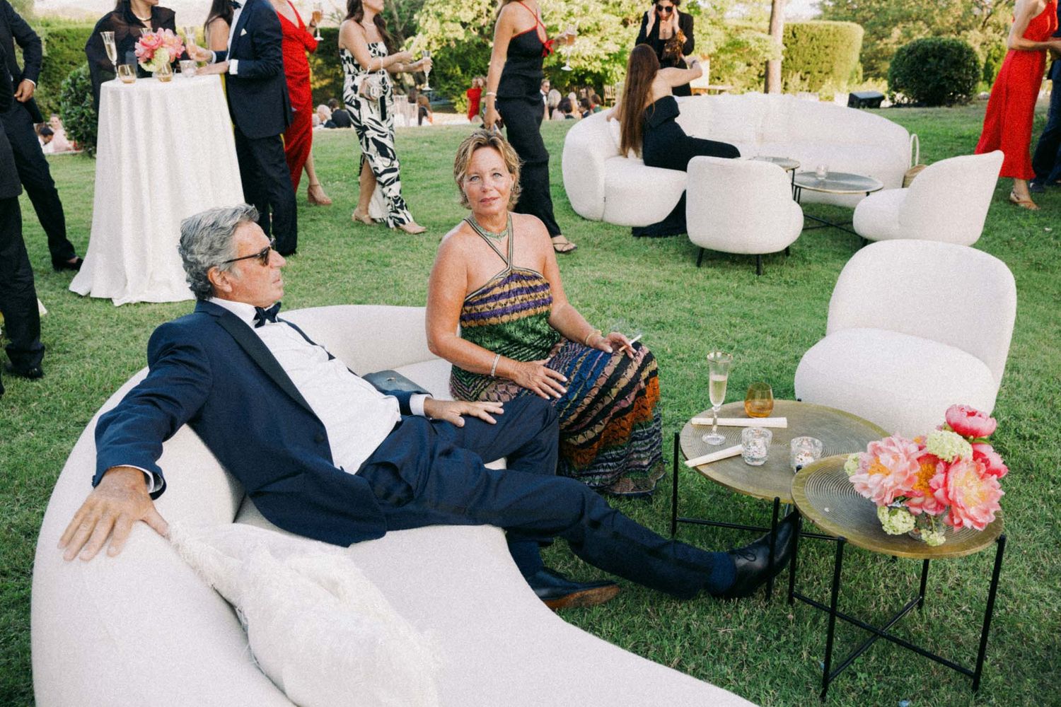 Guests mingle and relax on a grassy lawn at an elegant outdoor wedding reception with white decor.