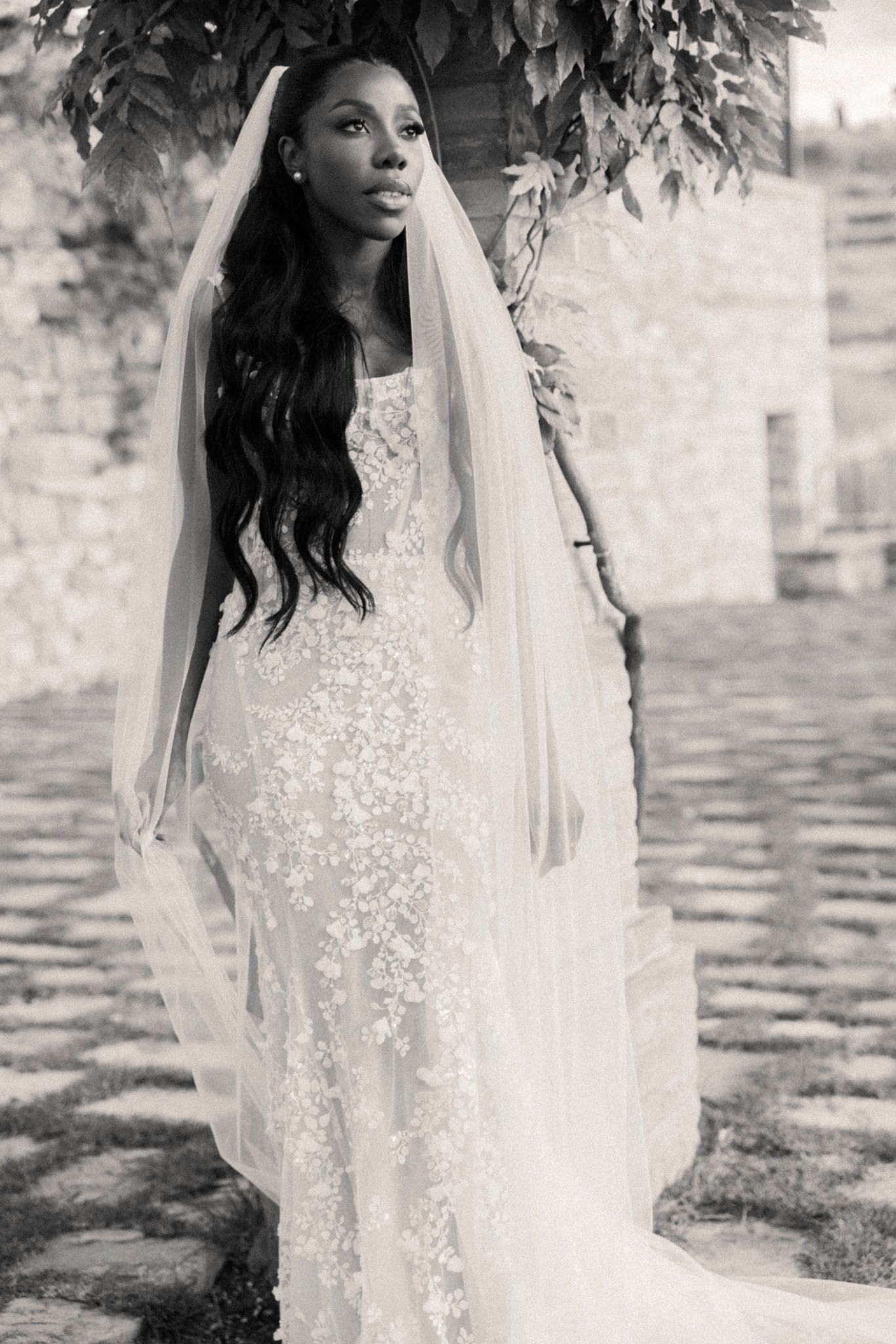 A bride in a delicate lace wedding dress poses gracefully in a garden setting, captured in black and white.
