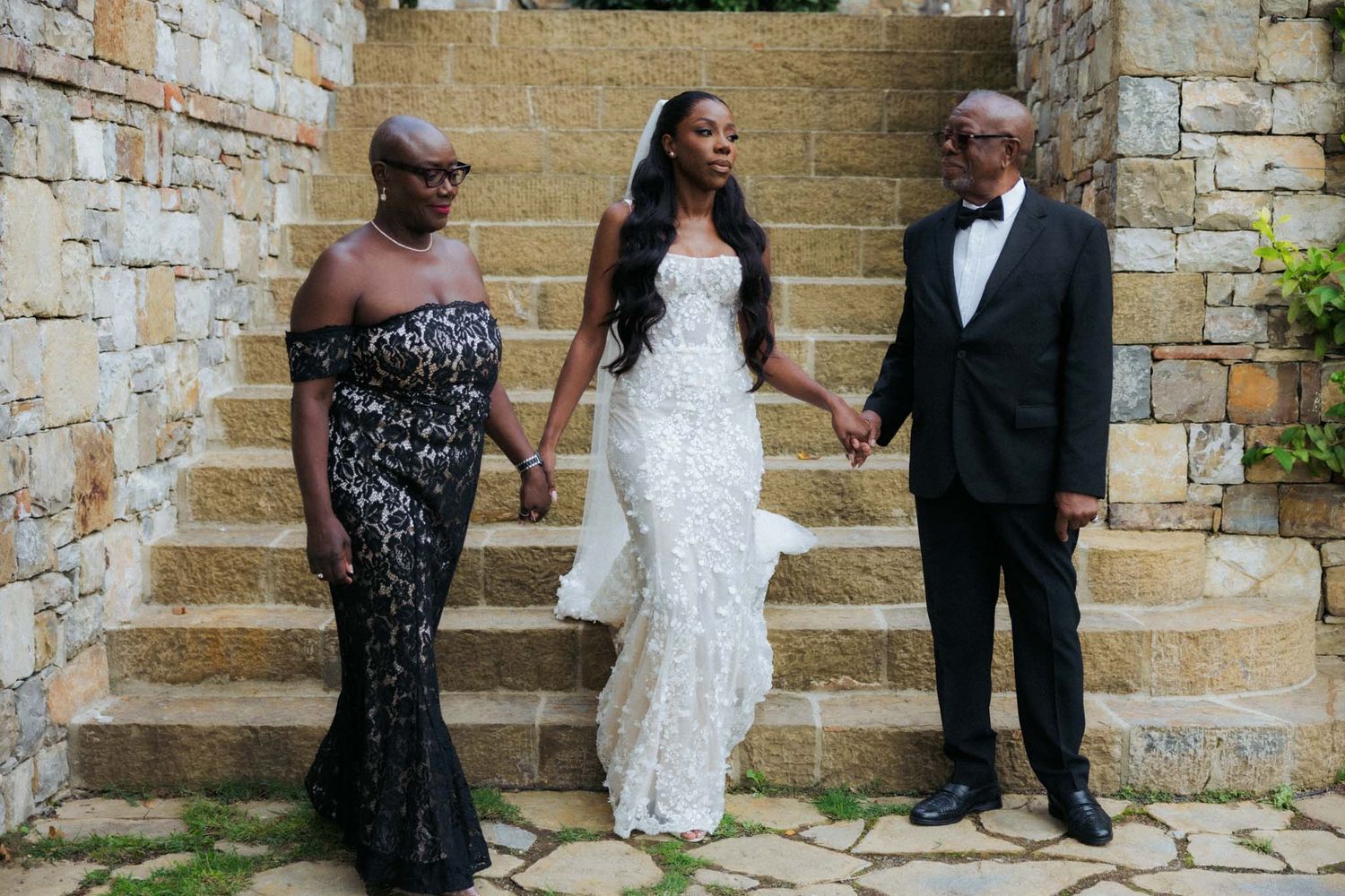 Wedding party members in formal attire pose against a rustic stone wall at an outdoor venue.