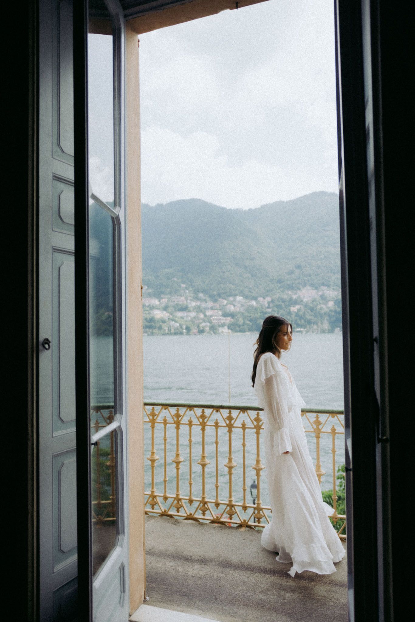 A person in a flowing white dress stands on a lakeside balcony with mountain views through an open doorway.