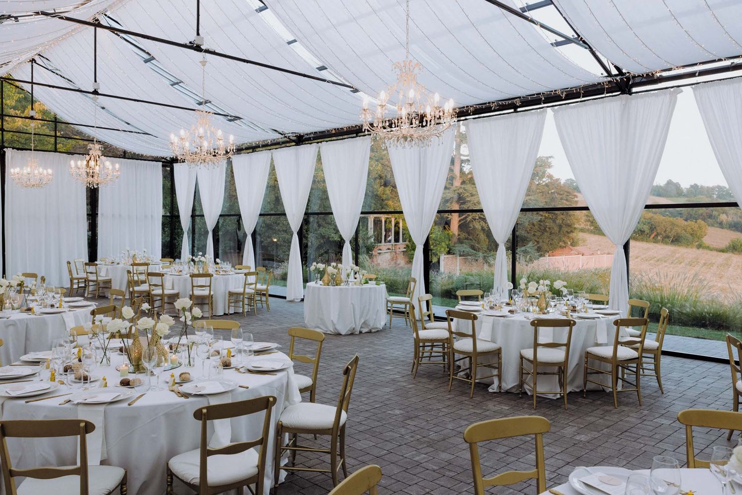 Wedding venue space decorated with white linens, elegant place settings, and crystal chandeliers overhead.