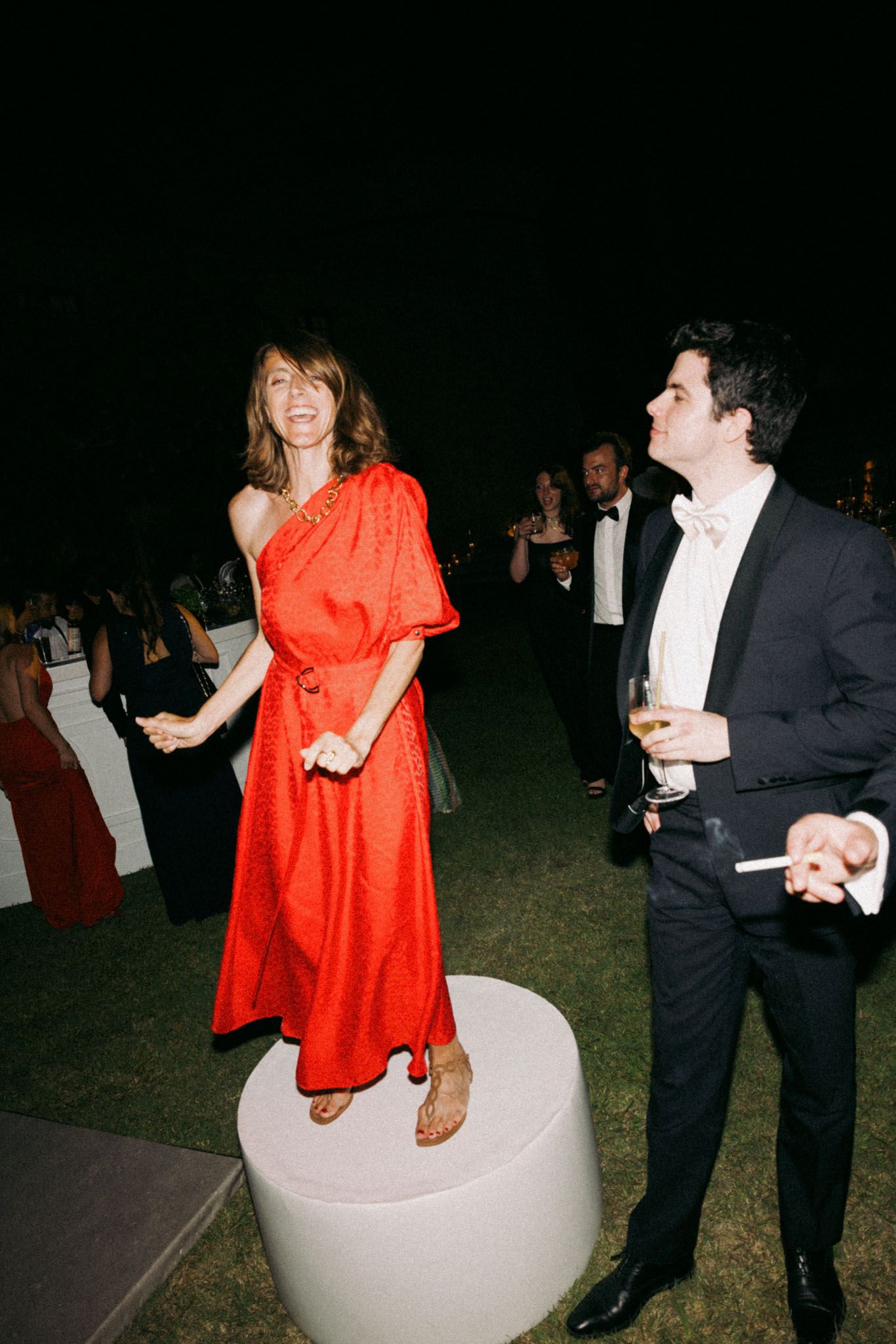 A person in a bright red dress sits on a white curved modern chair while conversing with someone in formal attire.