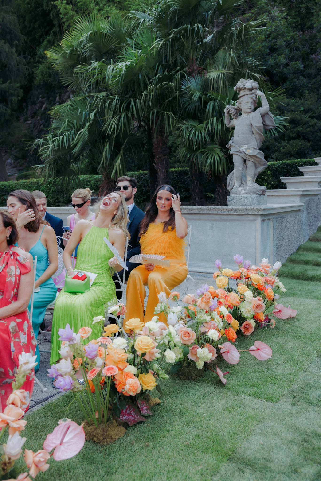 Bridal party in colorful floral dresses pose with cameras on a garden lawn with peach and pink flower arrangements.