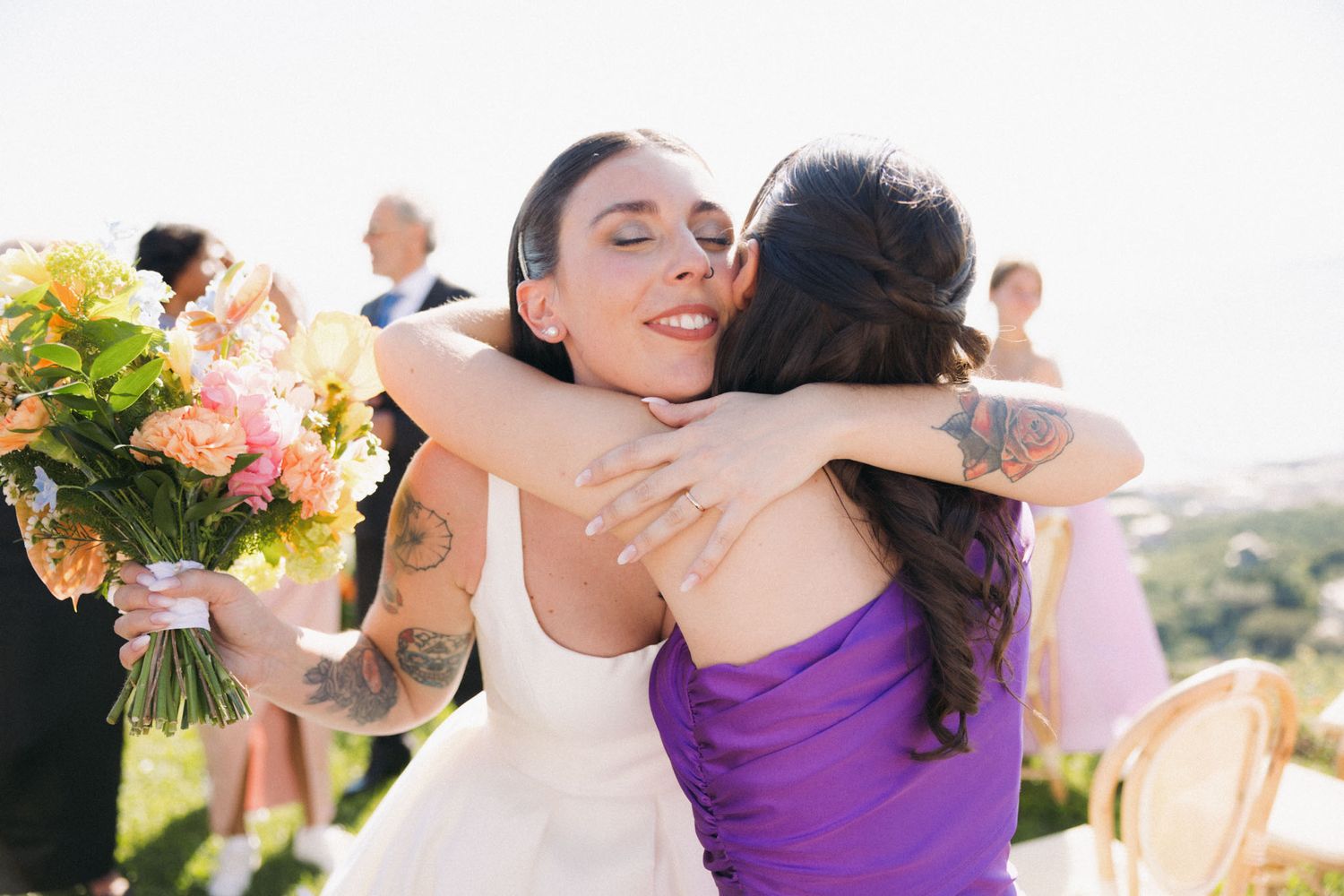 Two people embrace in a joyful hug at an outdoor wedding ceremony, with one holding a vibrant bouquet of flowers.