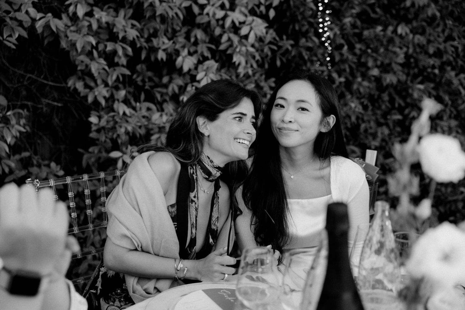 A black and white photo shows people gathered outdoors for a social event with drinks and conversation.