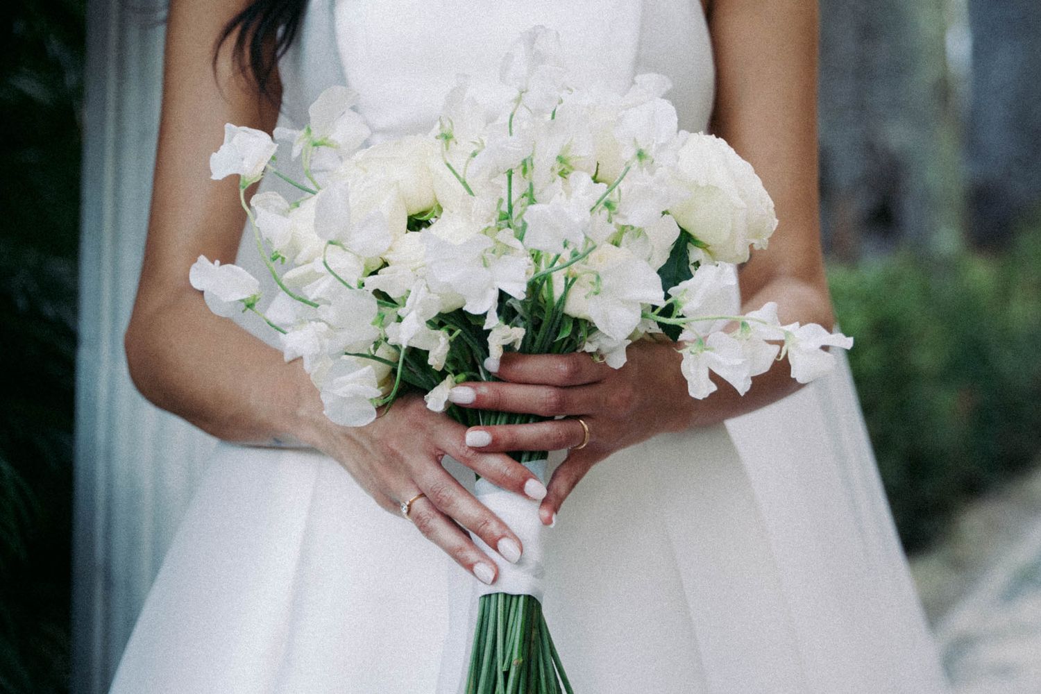 Close-up view of hands holding an elegant white floral wedding bouquet with roses and greenery against a white dress.