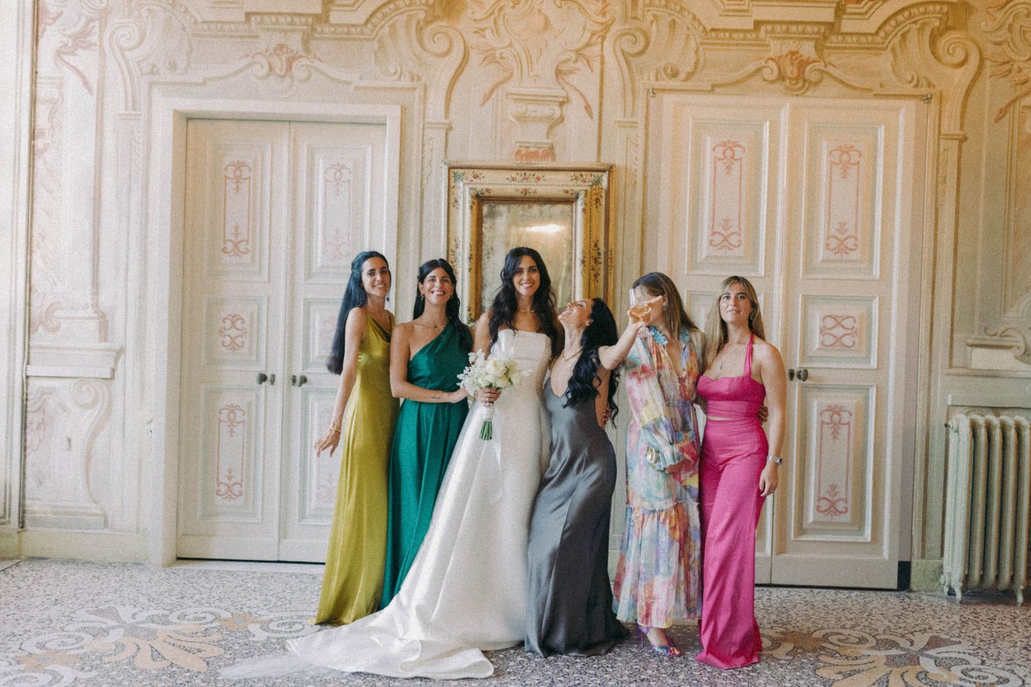Bridesmaids in colorful dresses line up against an ornate wall in an elegant wedding venue.