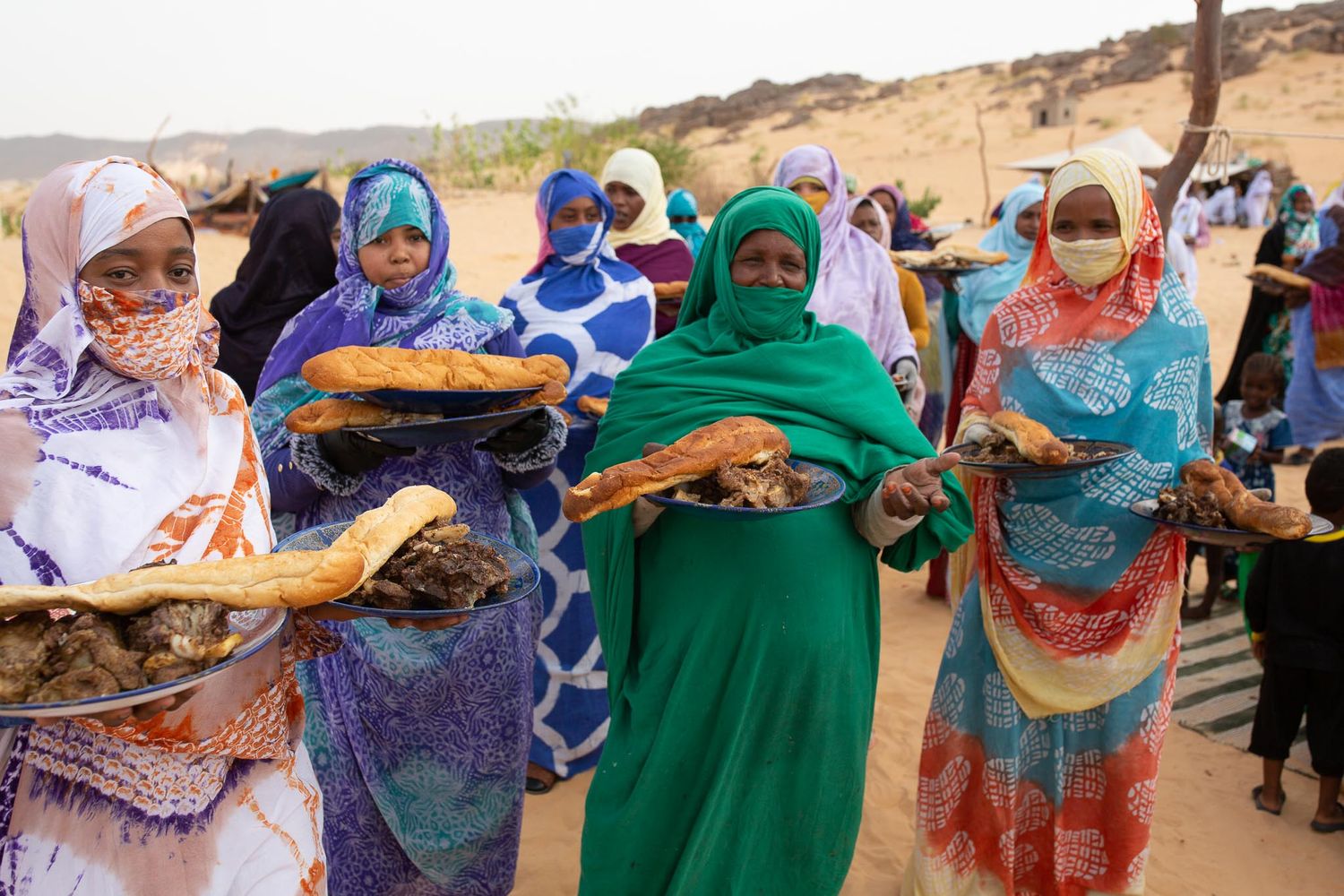 A group wearing colorful traditional garments gather outdoors holding textiles in vibrant patterns.