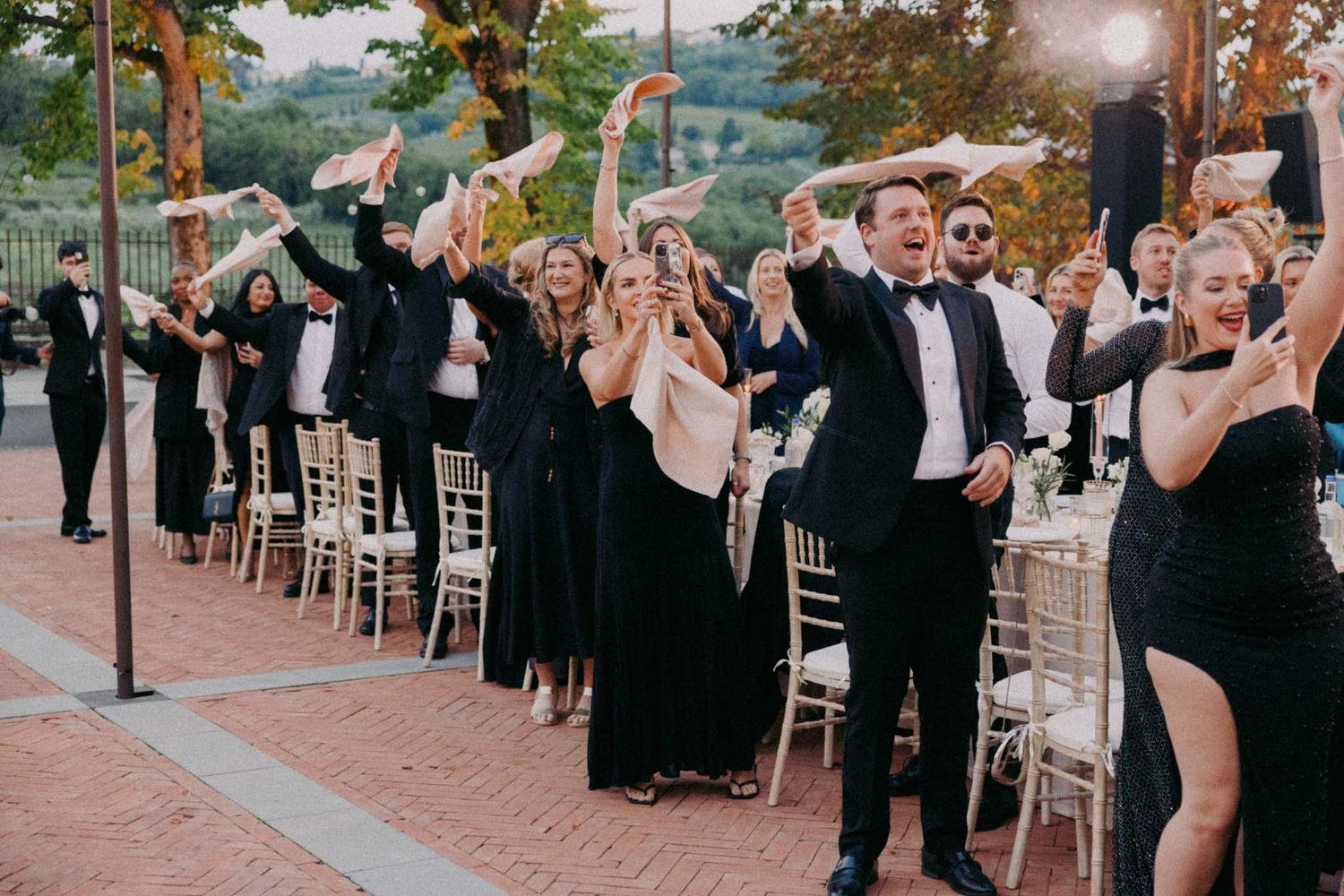 Wedding guests in formal black attire celebrate joyfully during an outdoor evening reception.