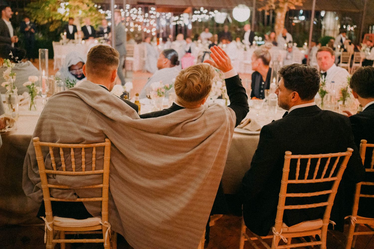 Wedding guests seated at tables with string lights overhead during an outdoor reception at sunset.