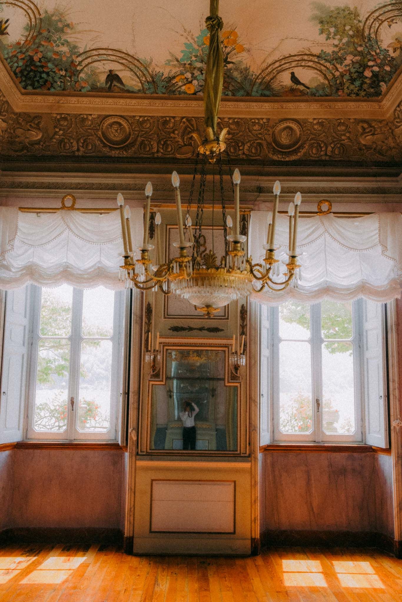 Panoramic view of an elegant historic room with ornate gold trim, chandeliers, and white curtained windows casting sunlight.