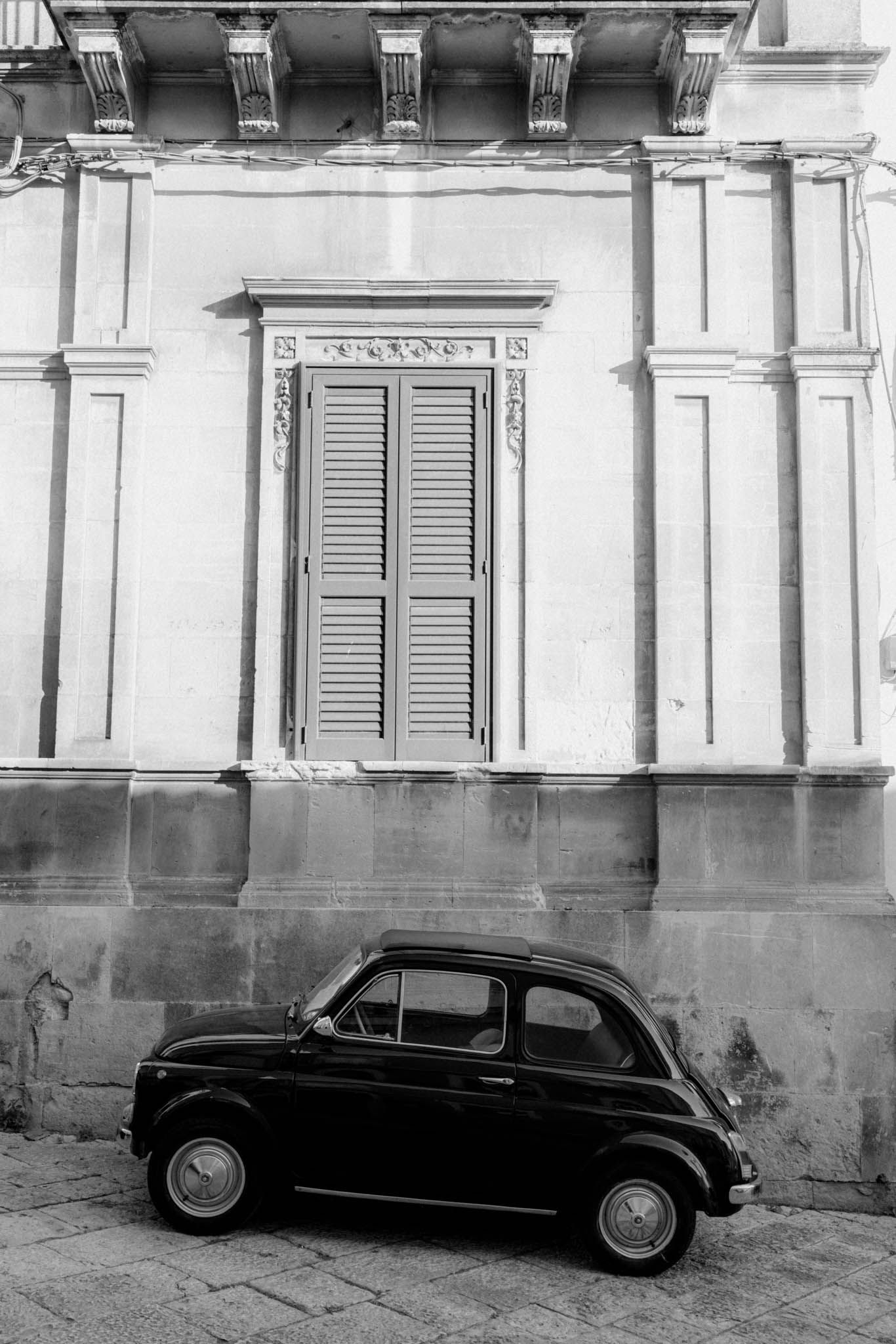 A vintage black car parked on a cobblestone street against a white building facade with shuttered windows.