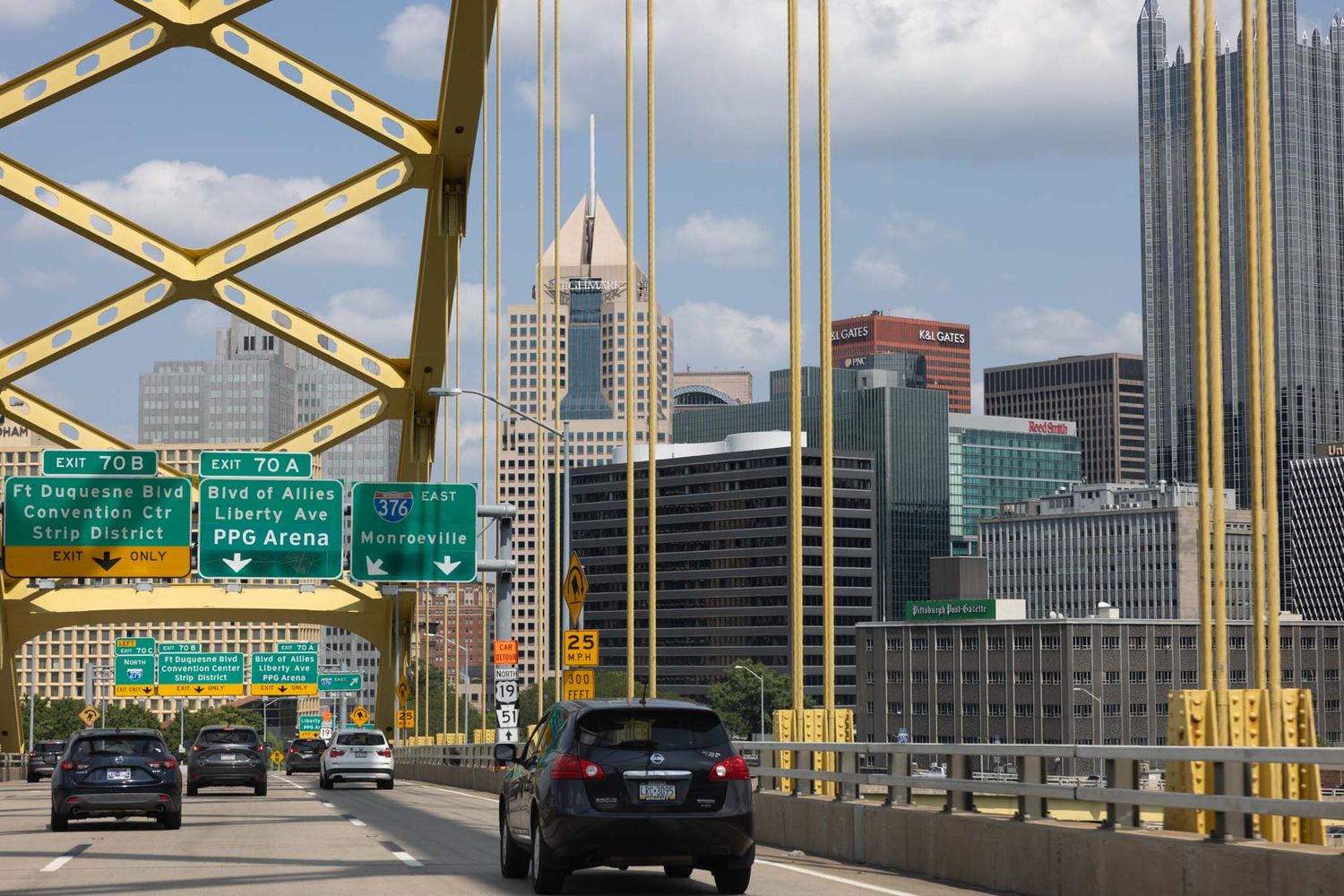 Cars drive across Pittsburgh's iconic yellow Fort Pitt Bridge with the downtown skyline visible in the background.
