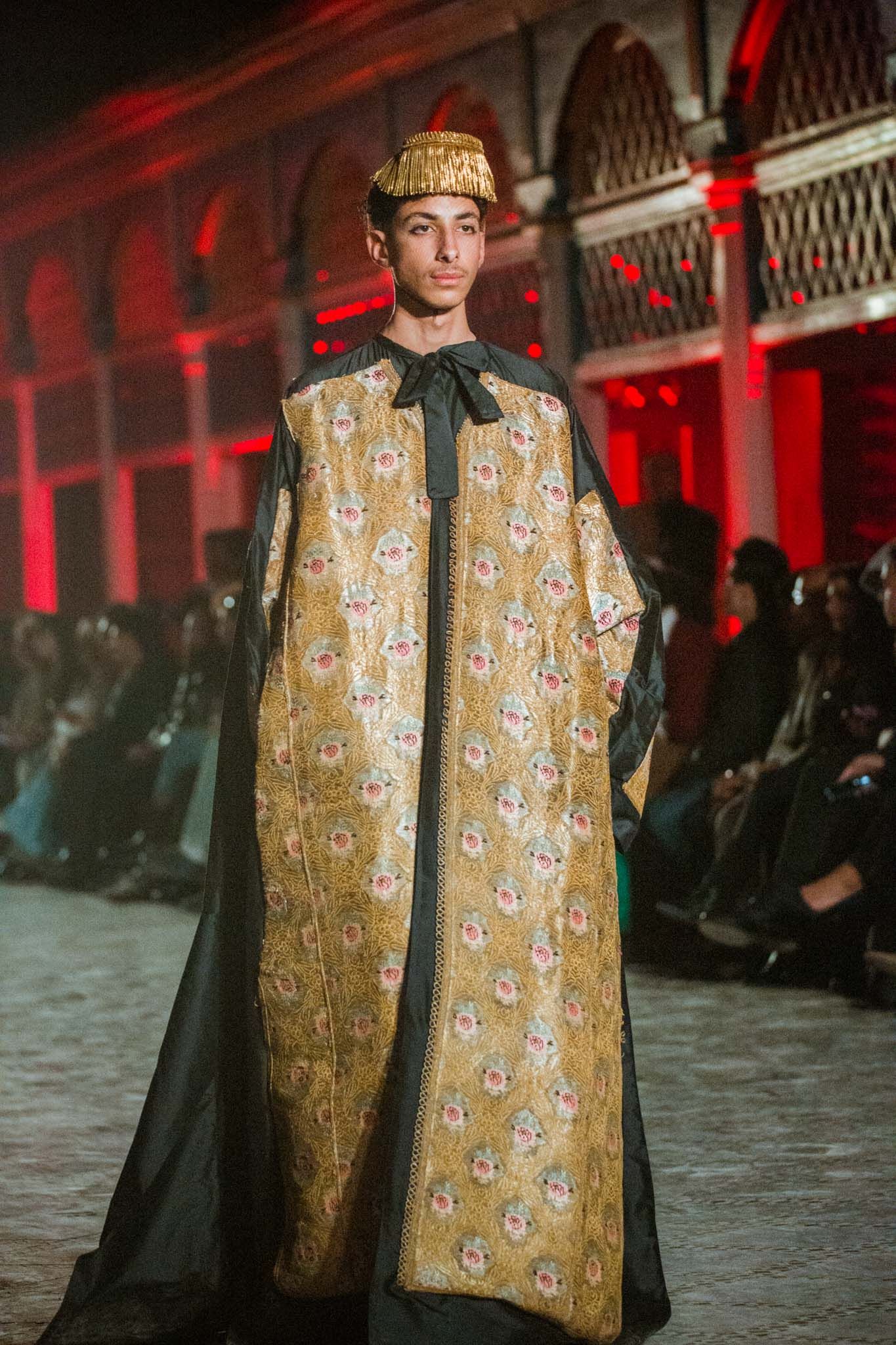 Model walks runway in gold floral brocade cape at fashion show with dramatic red lighting.