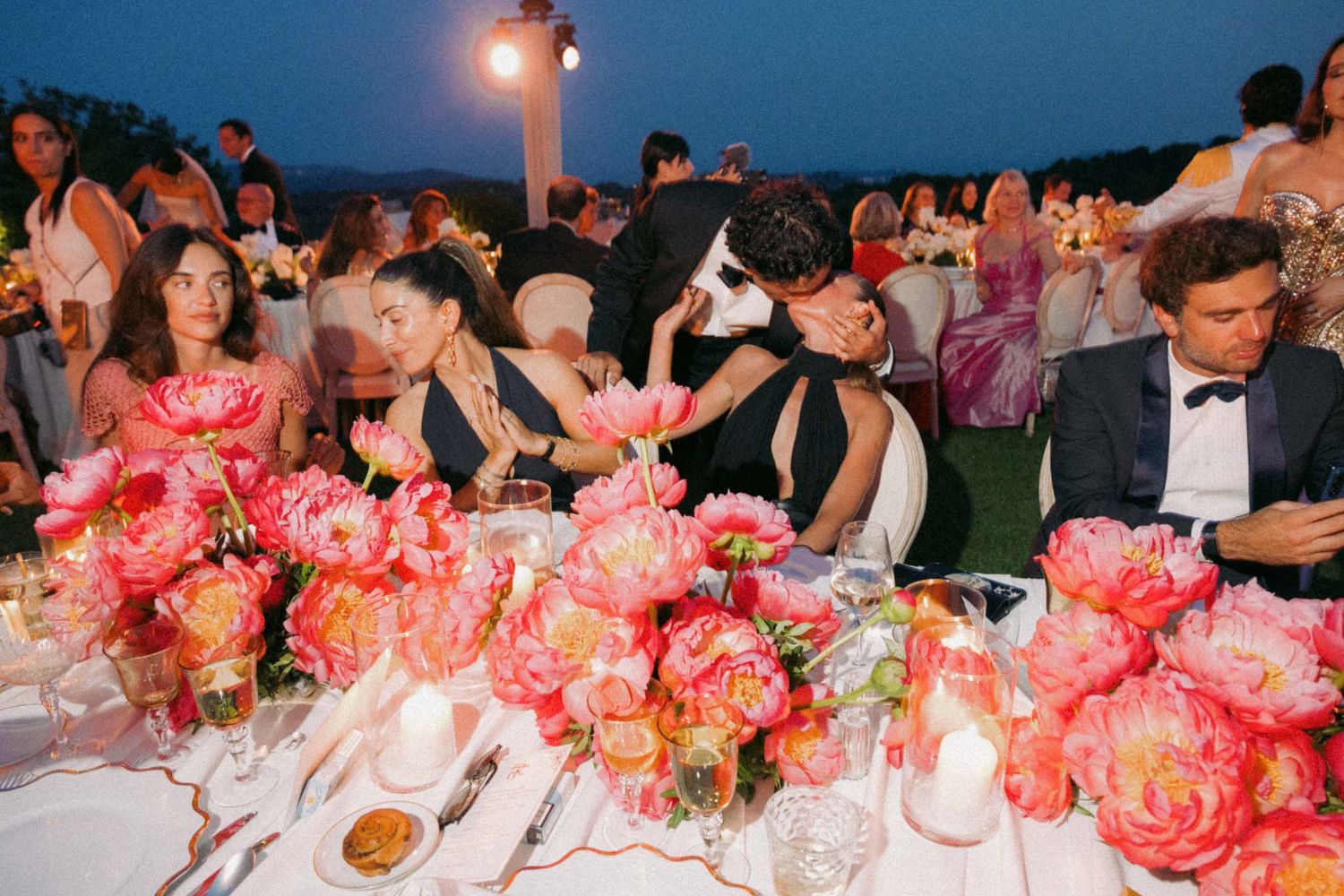 Wedding reception table decorated with vibrant coral and pink floral centerpieces glows under evening lighting.