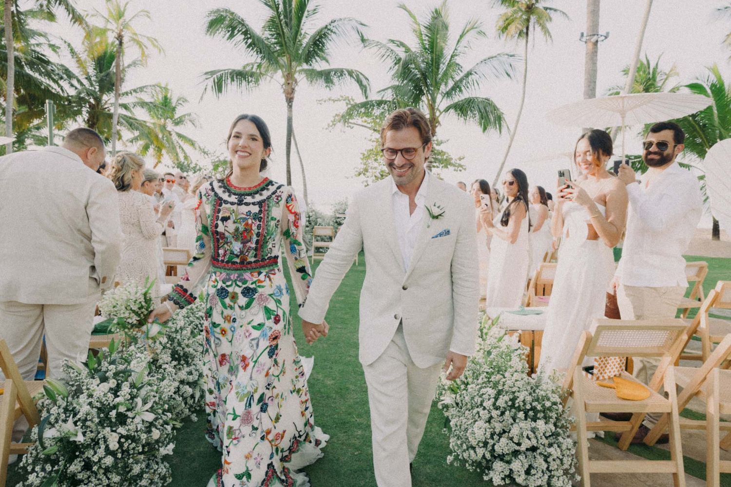 Guests celebrate at a tropical beach wedding reception with palm trees and white decor under a clear tent.