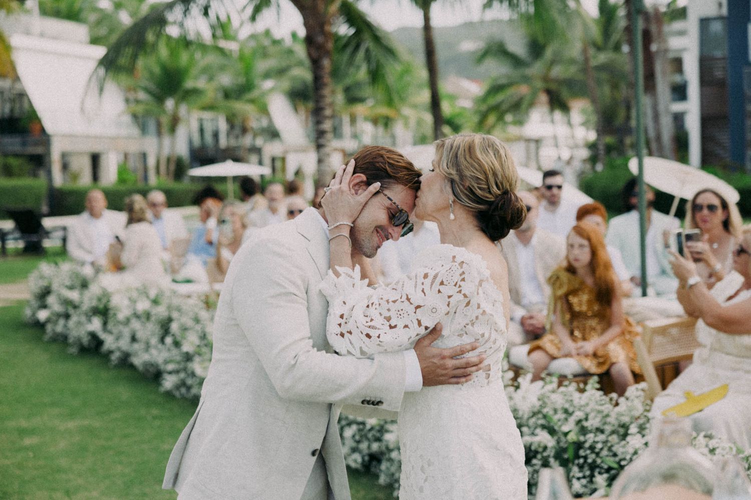 A romantic outdoor wedding celebration with a couple embracing on a lush green lawn surrounded by white floral decorations.