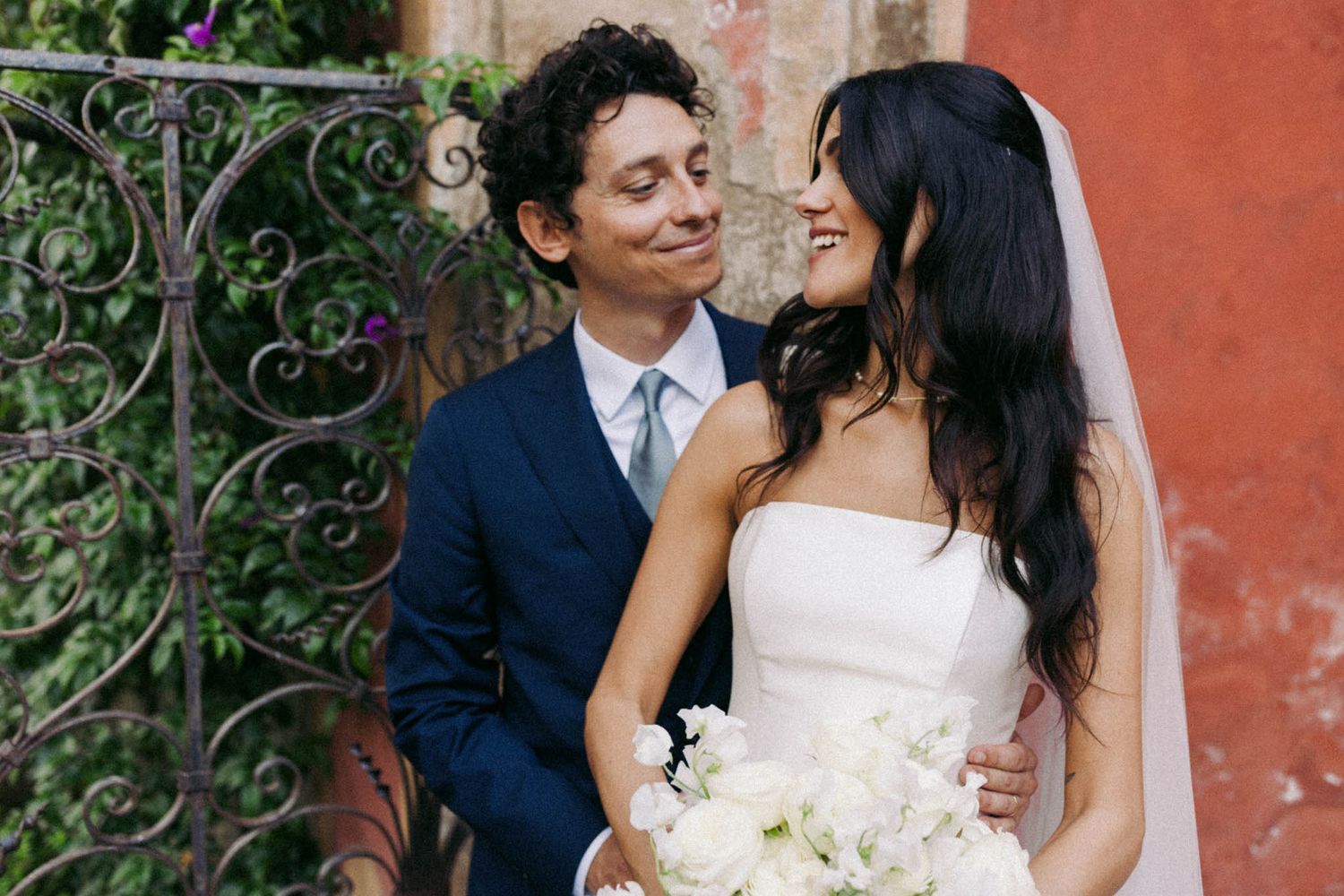 A couple shares a romantic moment in front of a rustic red wall with green foliage.