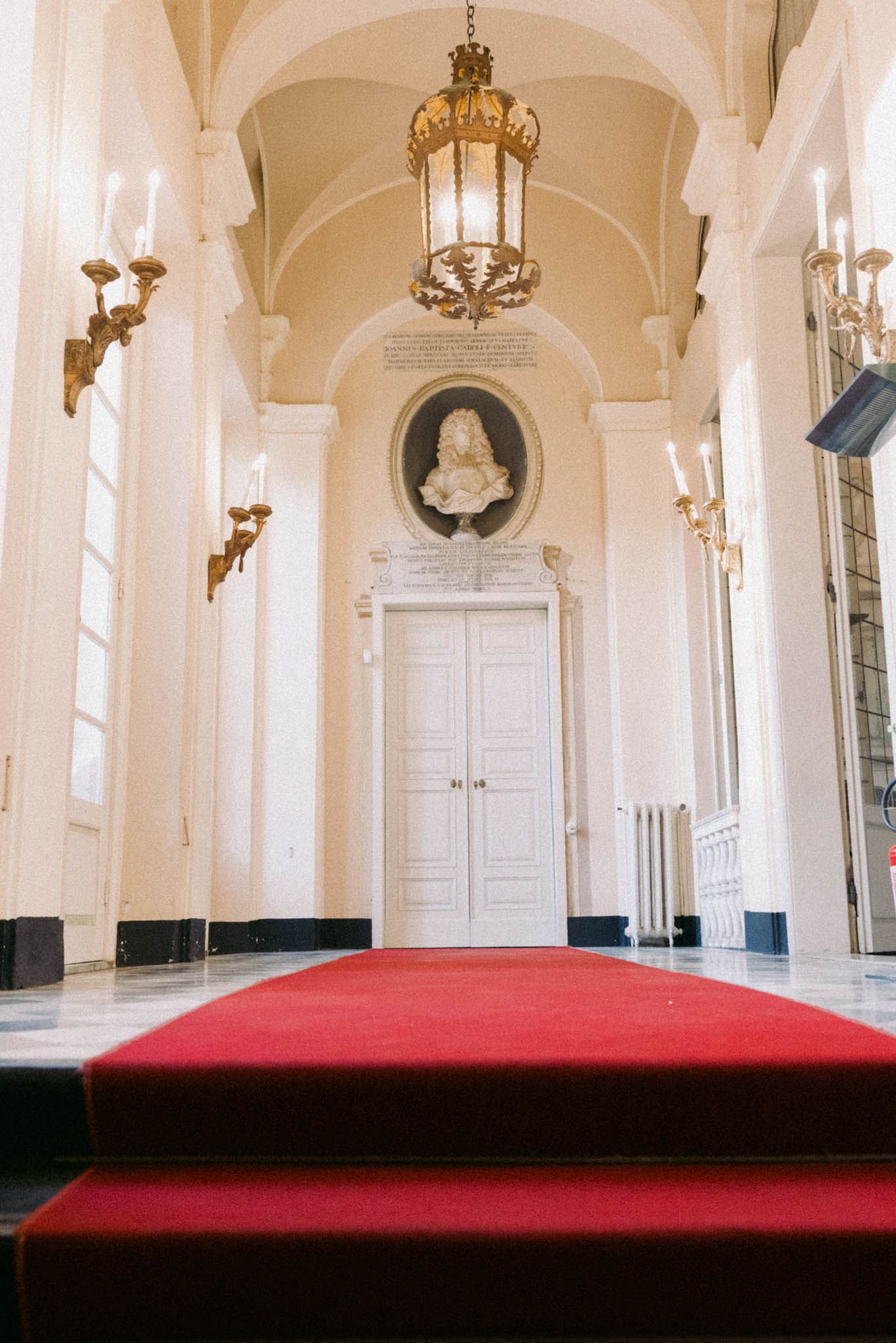 Elegant hallway with white arches, ornate brass chandelier, and red carpet steps leading to a grand white door.