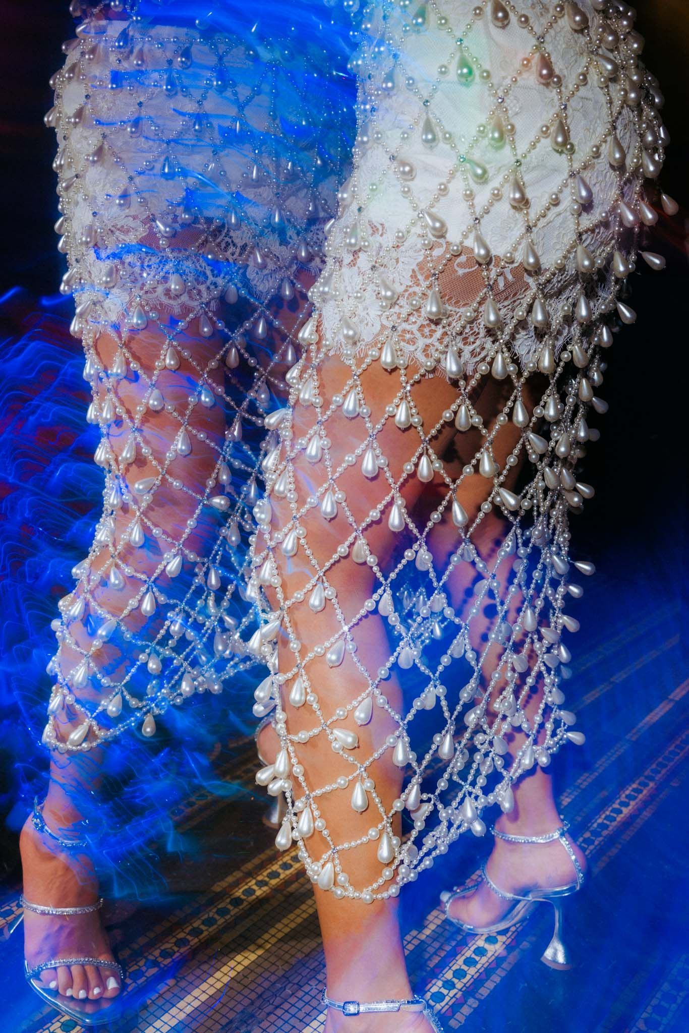 Close-up view of intricate beaded mesh dress detail with rhinestones glittering against a vibrant blue background.