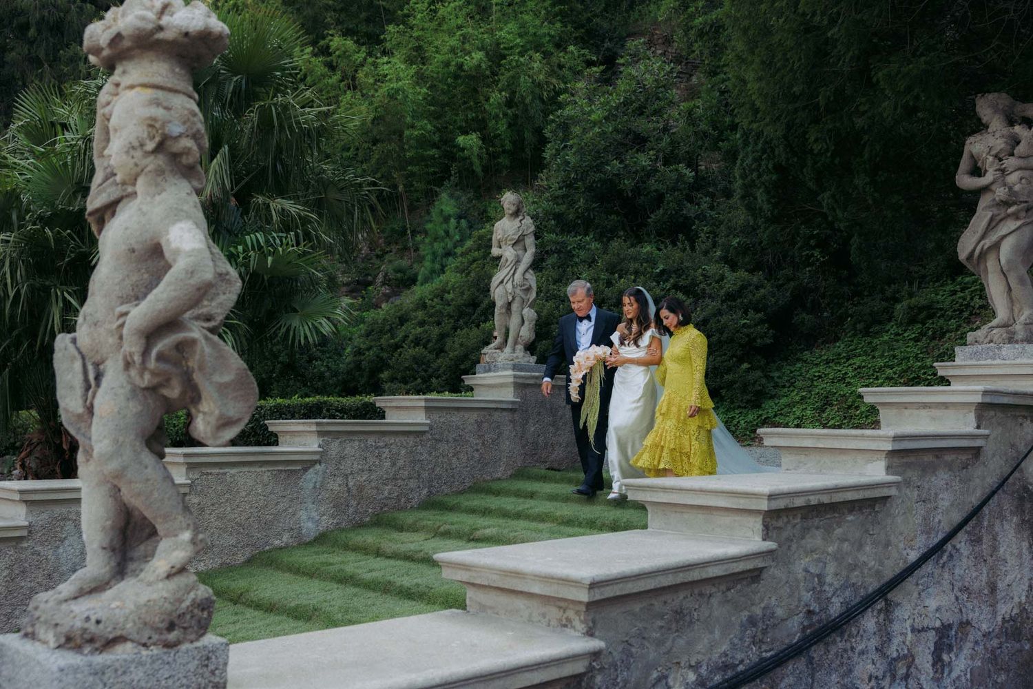 A collection of classical stone statues line a garden path with lush green foliage in the background.