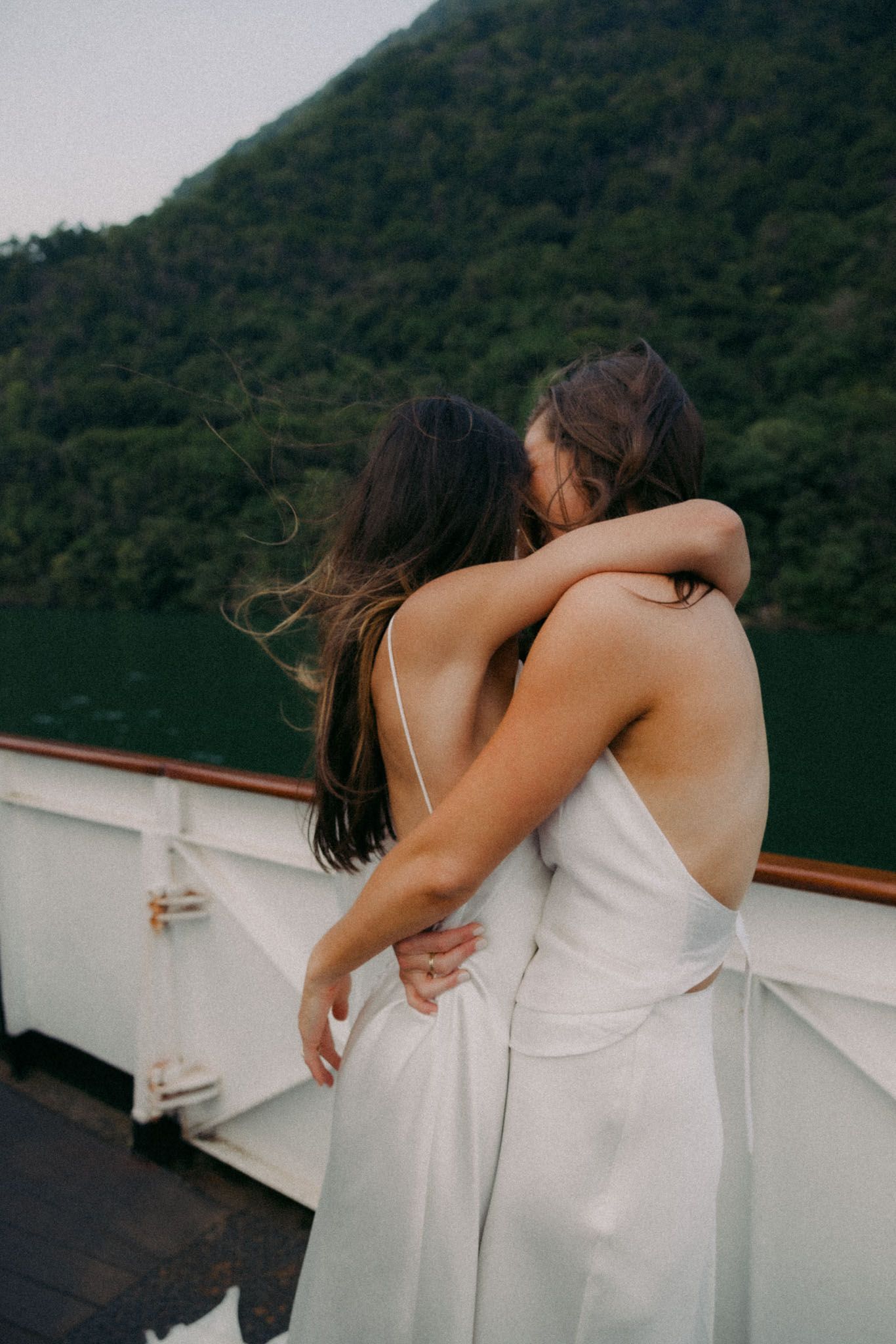 A series of candid moments showing two people in white dresses embracing on a boat with scenic mountains behind them.