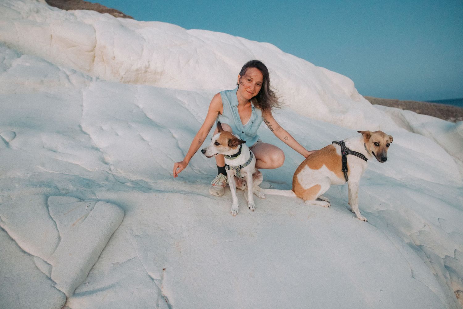A figure in blue poses with two dogs on white limestone rock formations against a bright turquoise sky.