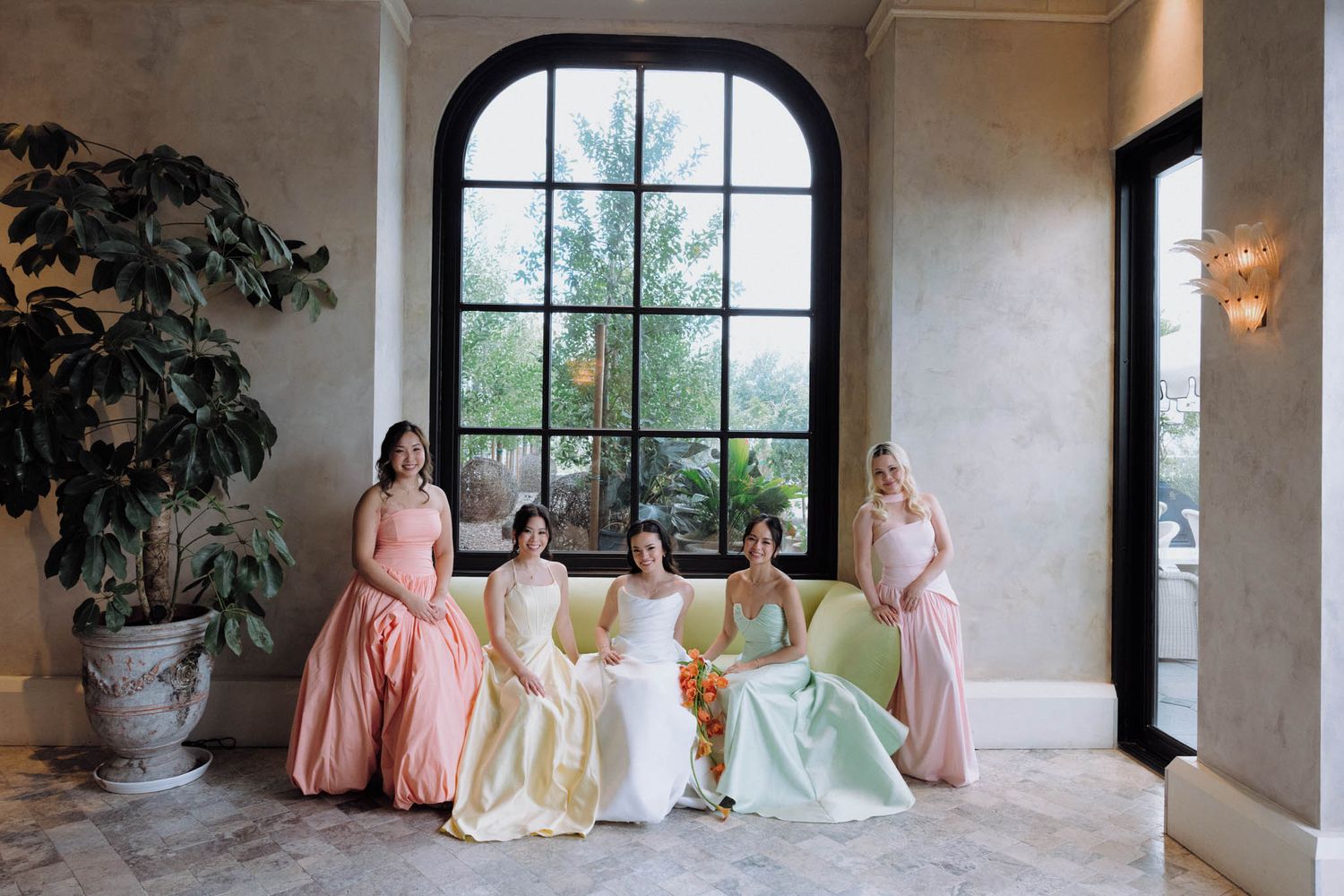 Bridal party in pastel dresses poses elegantly in a bright sunlit room with arched windows and stone walls.