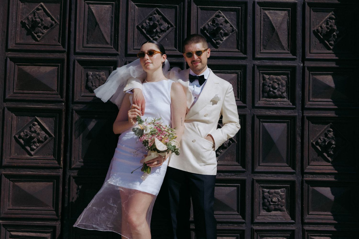 A couple in formal wedding attire share a romantic moment against an ornate dark wooden door background.