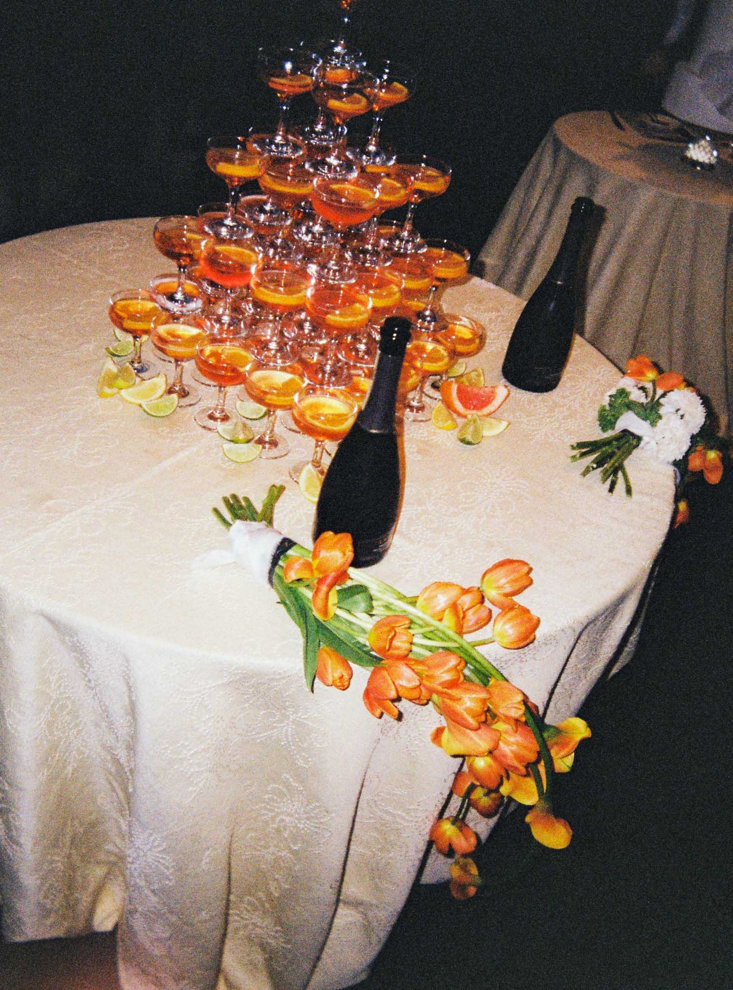 A champagne tower with orange cocktails sits on a white tablecloth next to orange tropical flowers.
