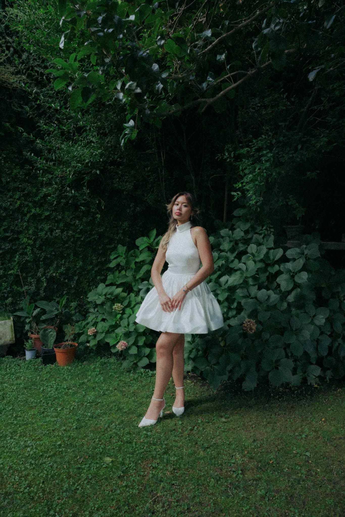 A figure in a white summer dress sits thoughtfully on grass surrounded by lush green foliage in a garden setting.
