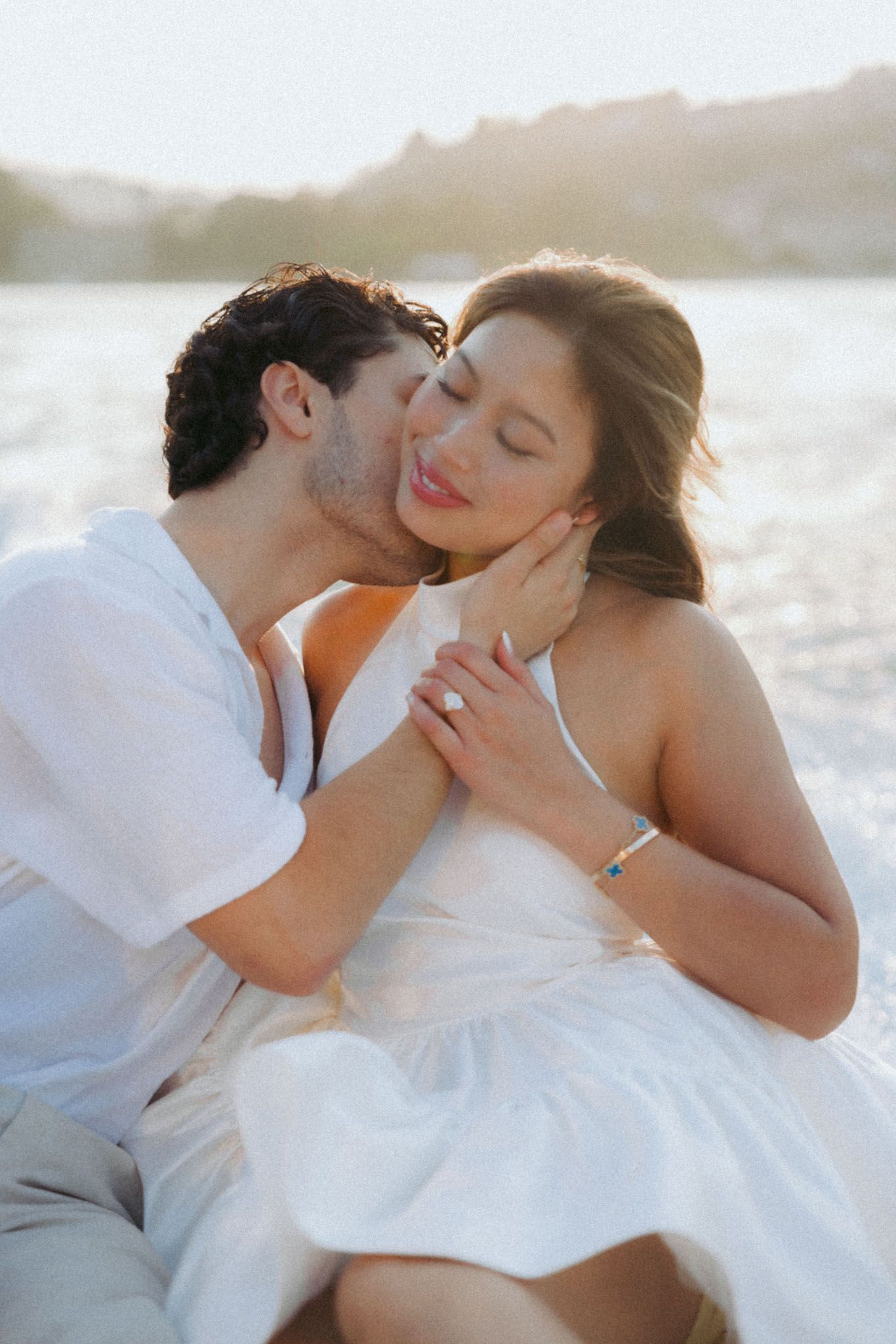 A romantic couple in white attire shares intimate moments on a sunlit beach during a photo session.