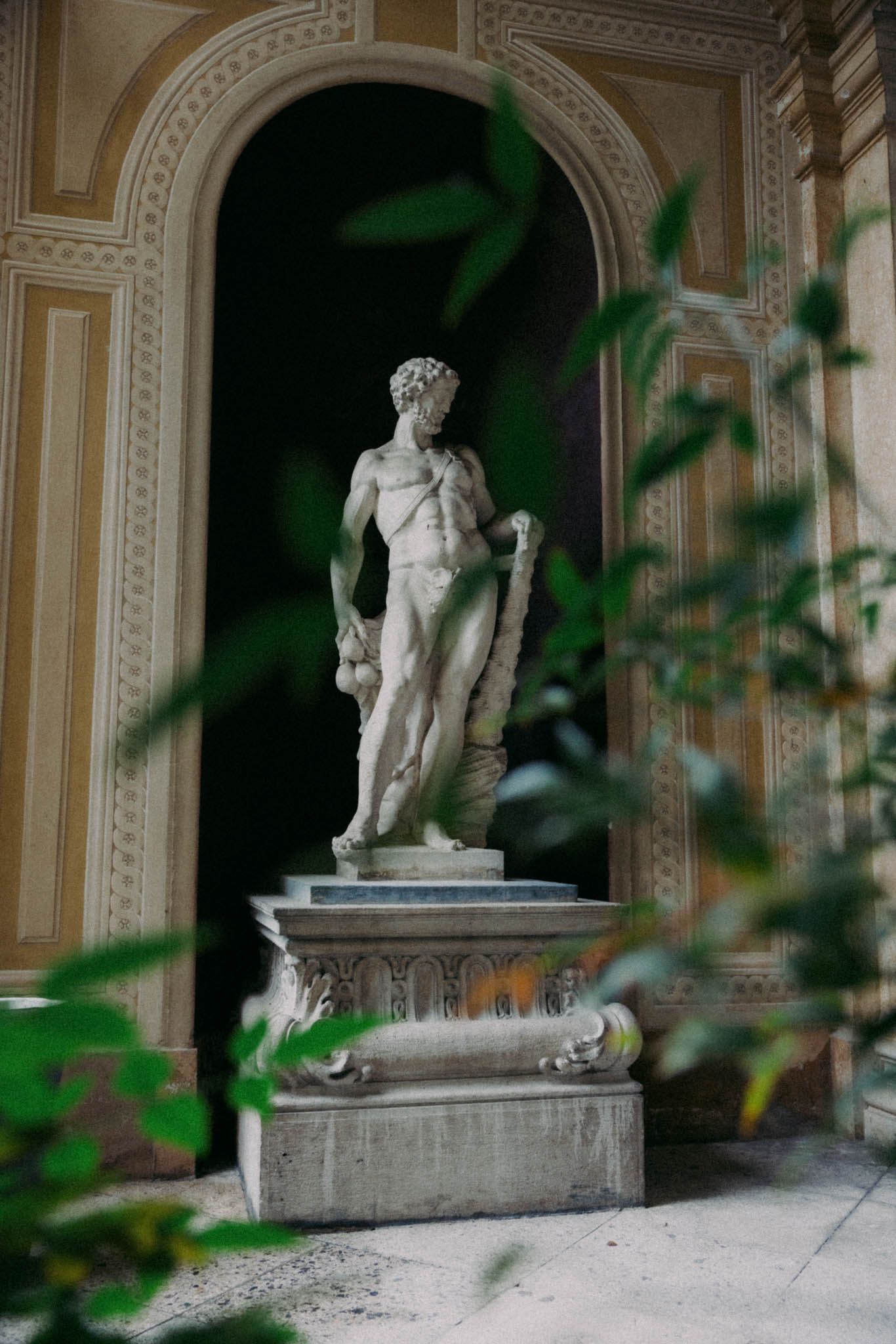 Classical white marble statues stand in ornate niches along a corridor with green foliage in the foreground.