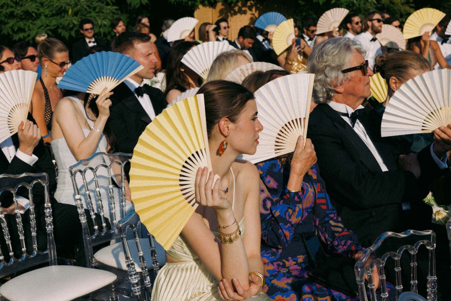 Wedding guests use paper fans to stay cool during an outdoor summer ceremony.