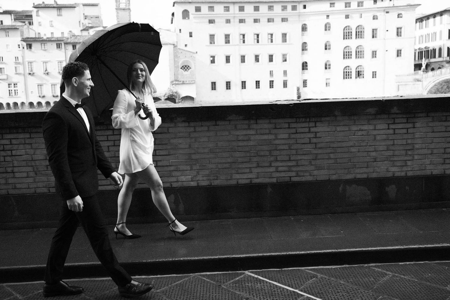 Two people walk with umbrellas along a terrace overlooking Italian buildings in black and white photography.