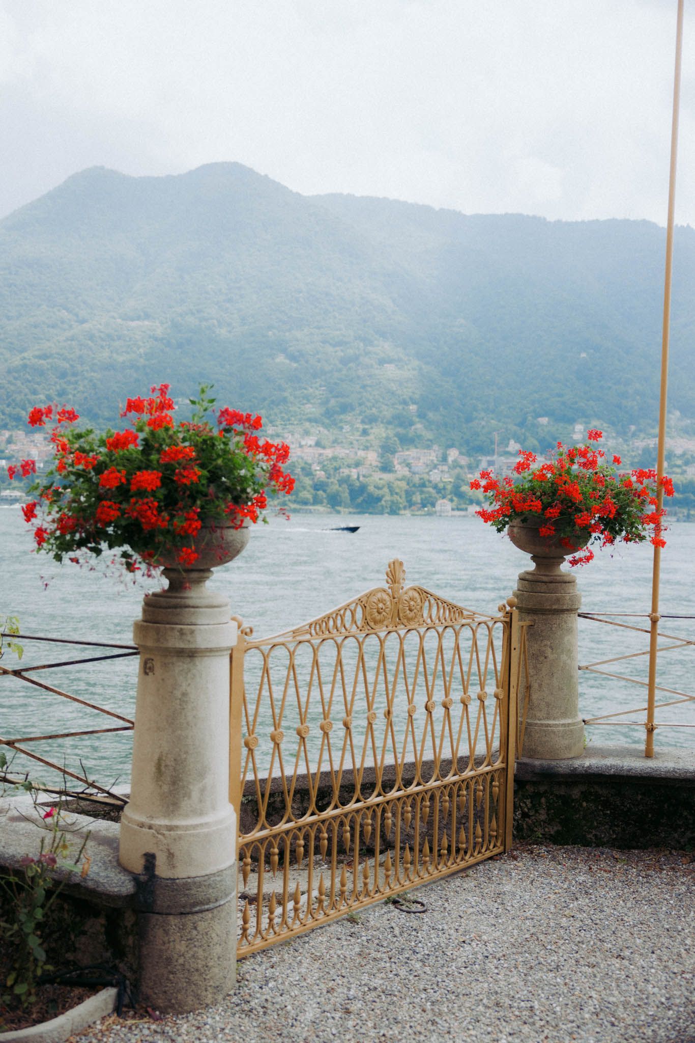 Ornate stone balcony with red geranium planters overlooking Lake Como and misty Italian mountains in the distance.