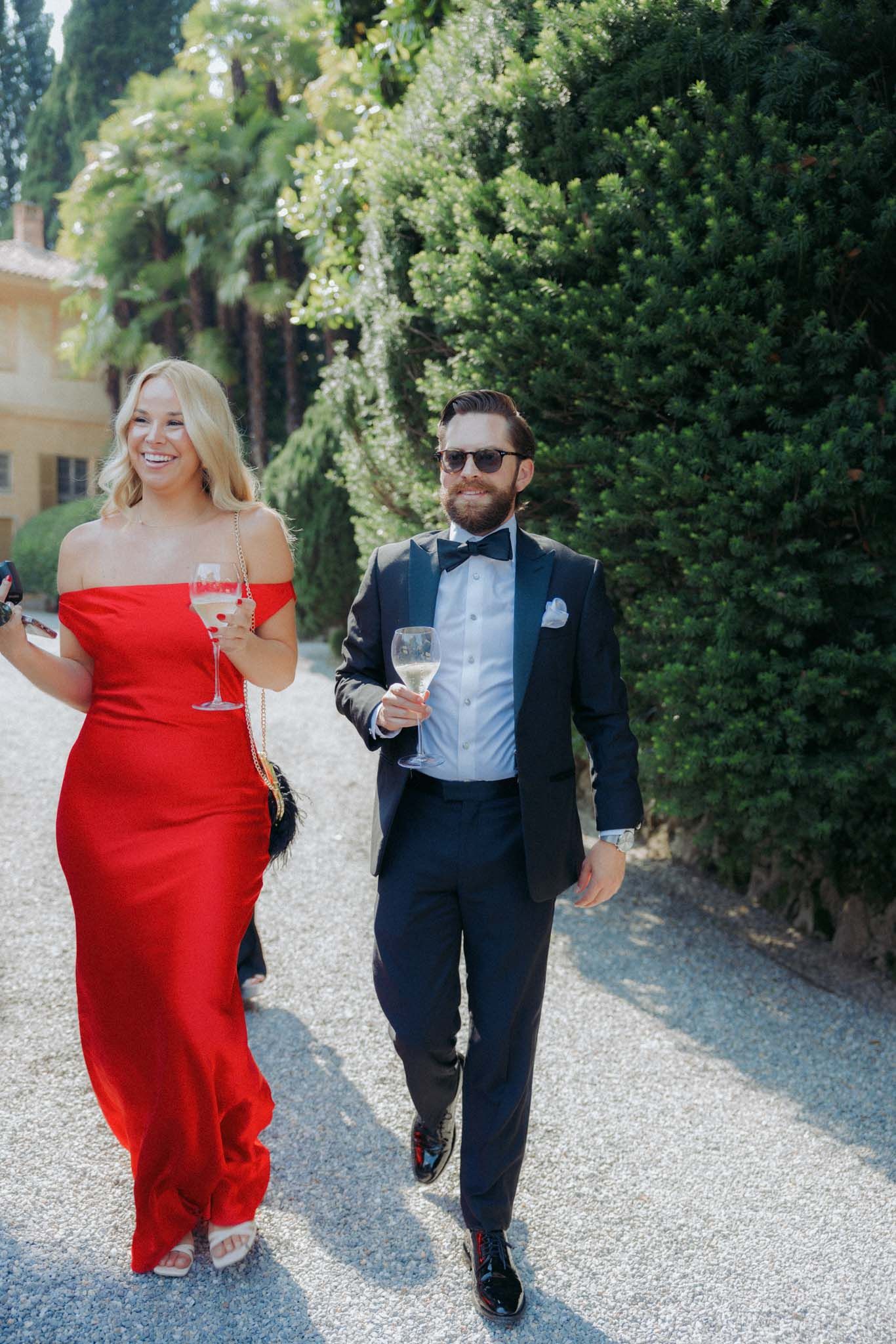 A couple in formal attire walks together on a gravel path surrounded by lush greenery.