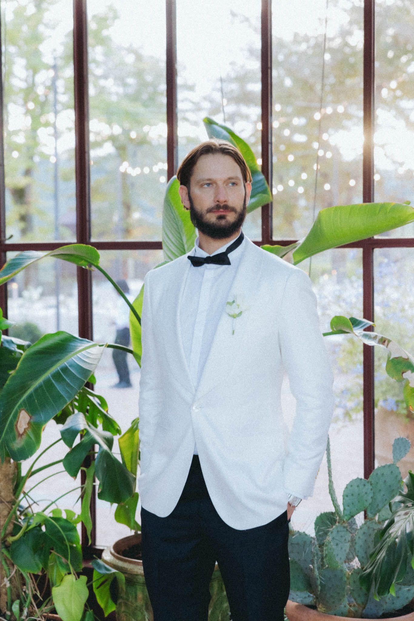 Two people in white shirts and dark vests stand in a greenhouse with lush green plants and natural lighting.