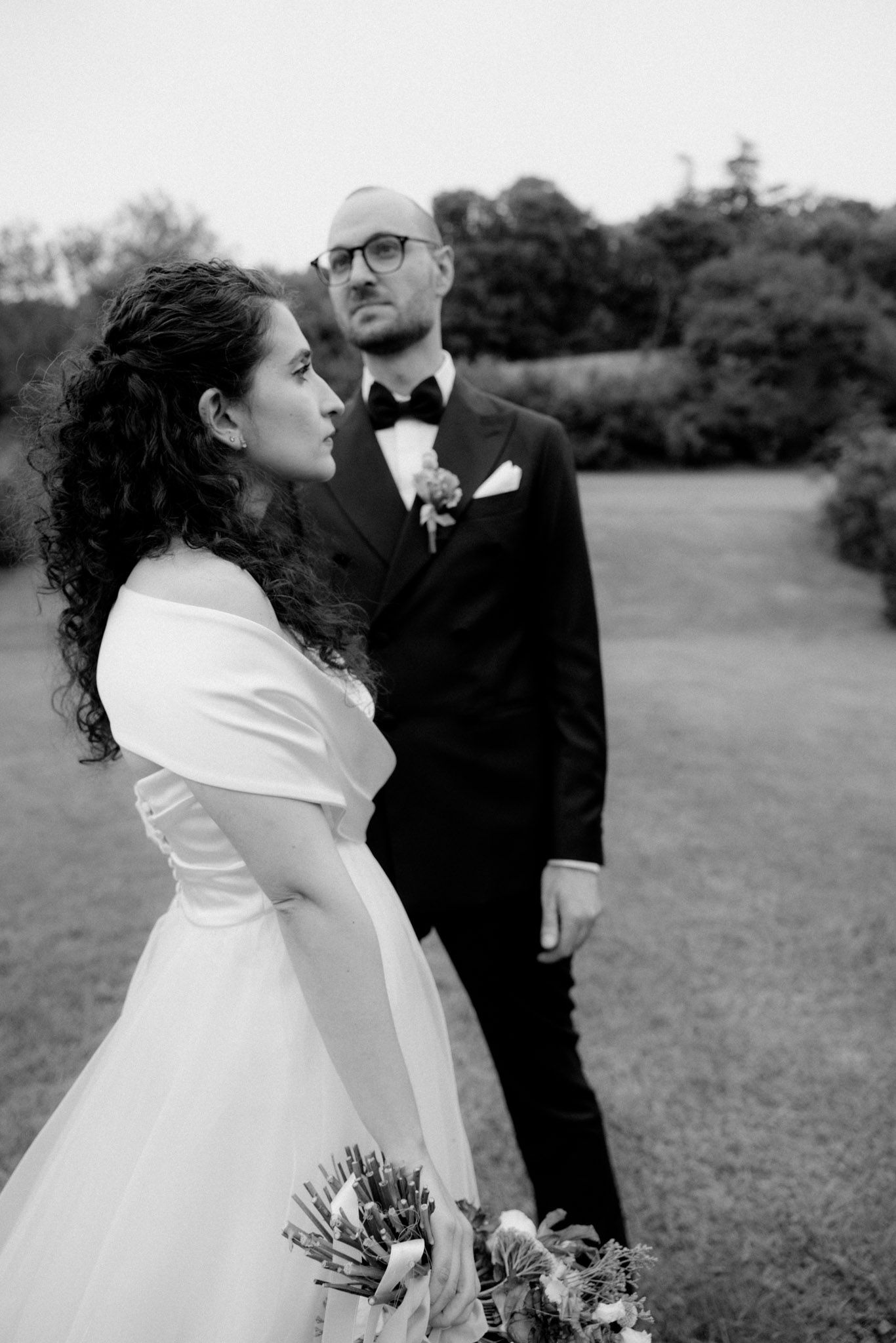 A black and white wedding portrait showing a bride with curly hair holding a bouquet beside her groom in a tuxedo.