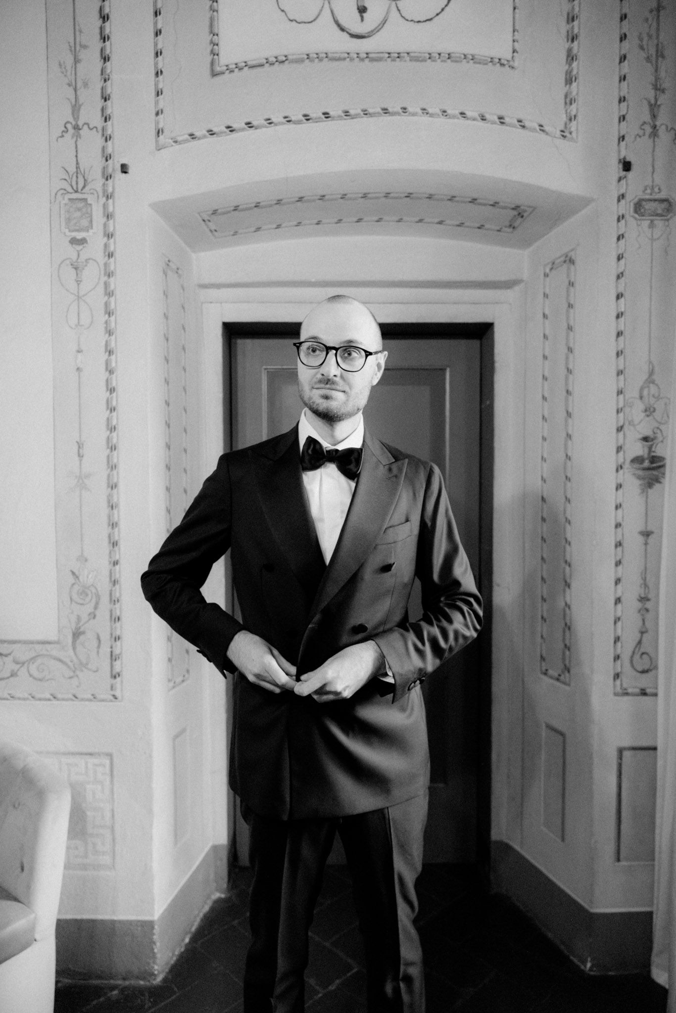 Black and white photograph showing a man in a formal tuxedo and bow tie standing in an ornate hallway.