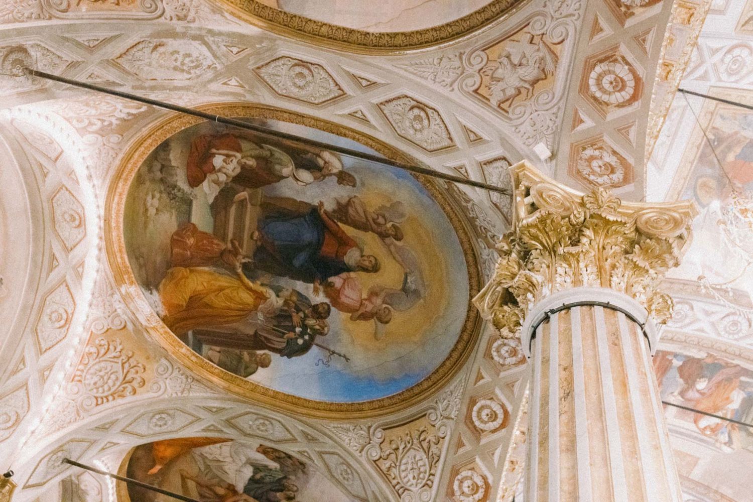 Ornate baroque ceiling fresco with religious figures surrounded by elaborate gilded architectural details and columns.