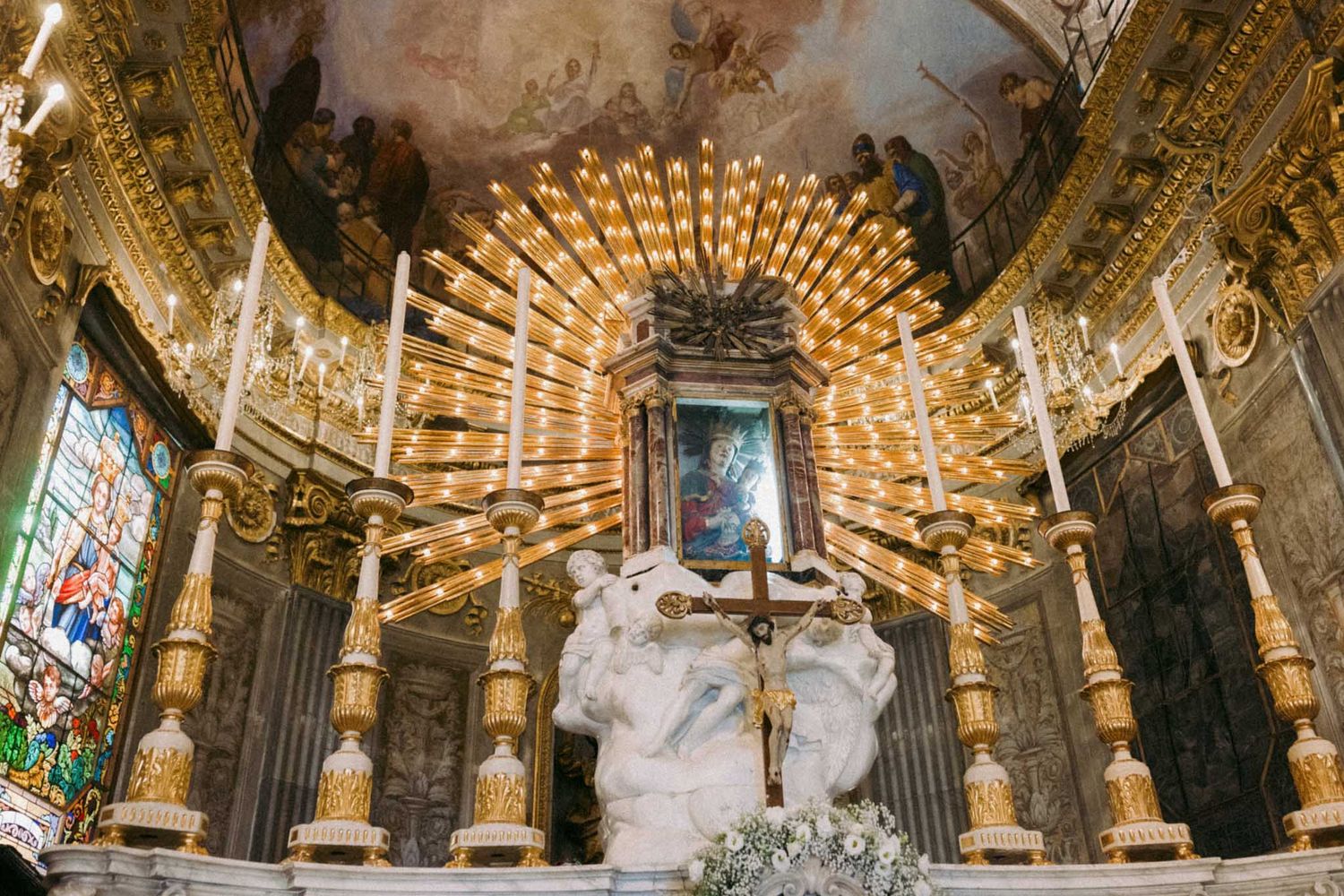 Ornate gold and crystal chandeliers illuminate the luxurious interior of a baroque palace with painted ceiling frescoes.