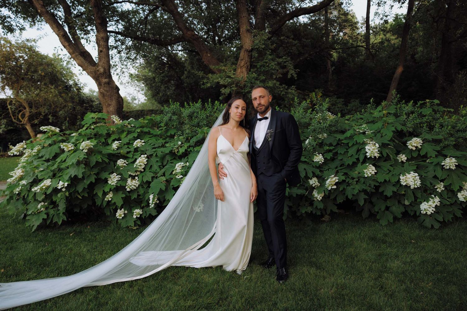 Wedding couple portraits in a lush garden setting with tall trees and white flowers.