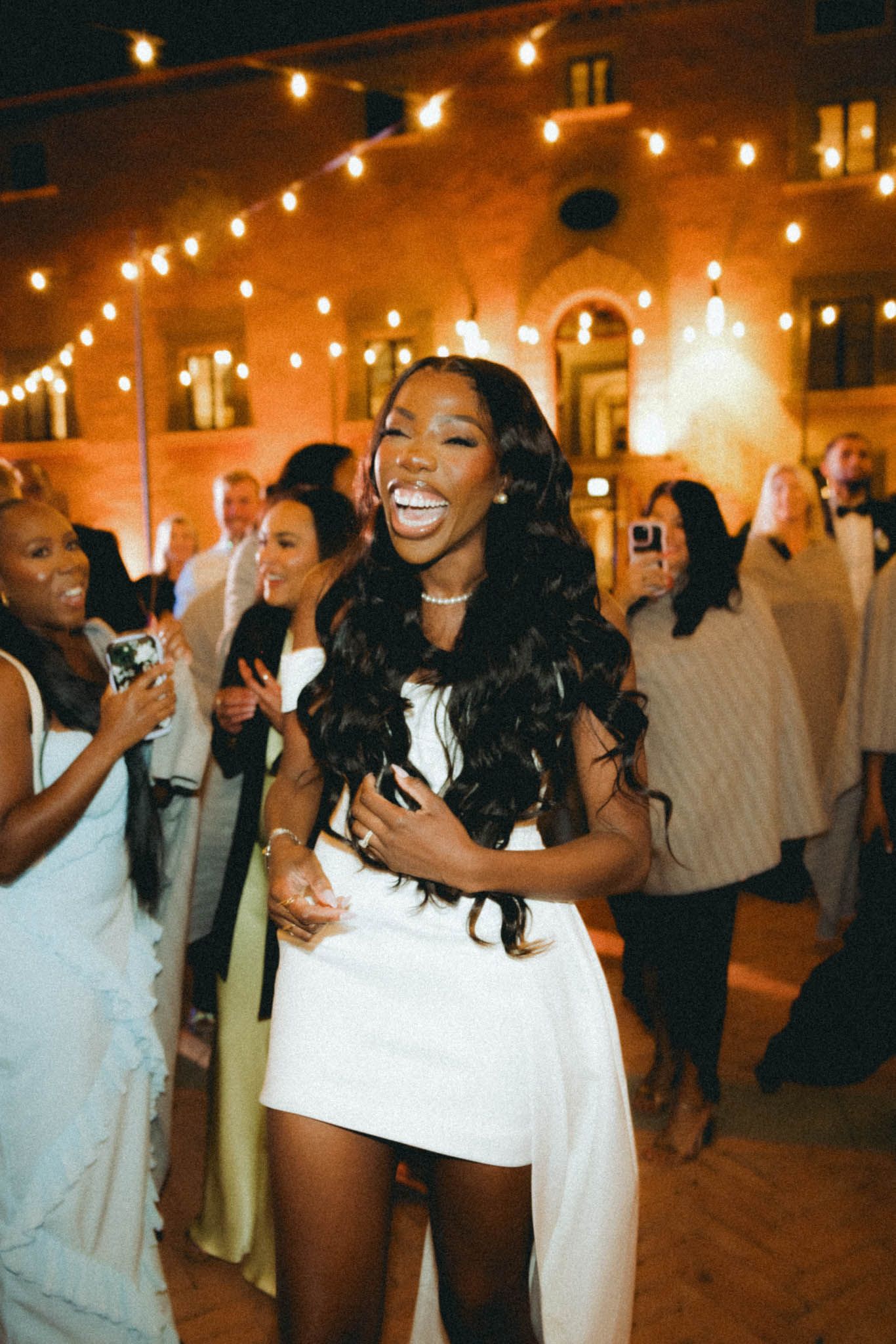 A joyful celebration on a dance floor illuminated by warm string lights at an indoor reception venue.