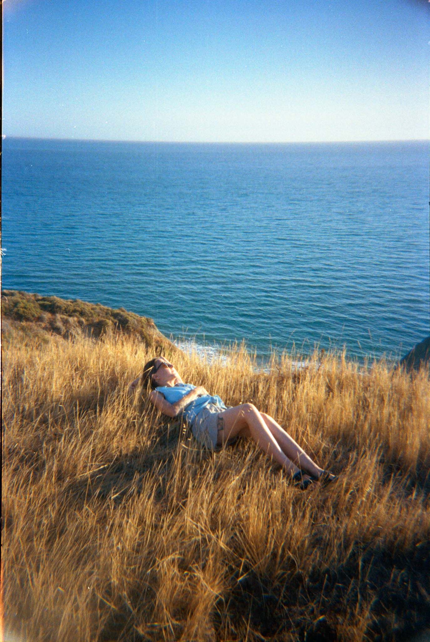 Person lying in golden grass on coastal cliff overlooking calm blue ocean waters on sunny day.