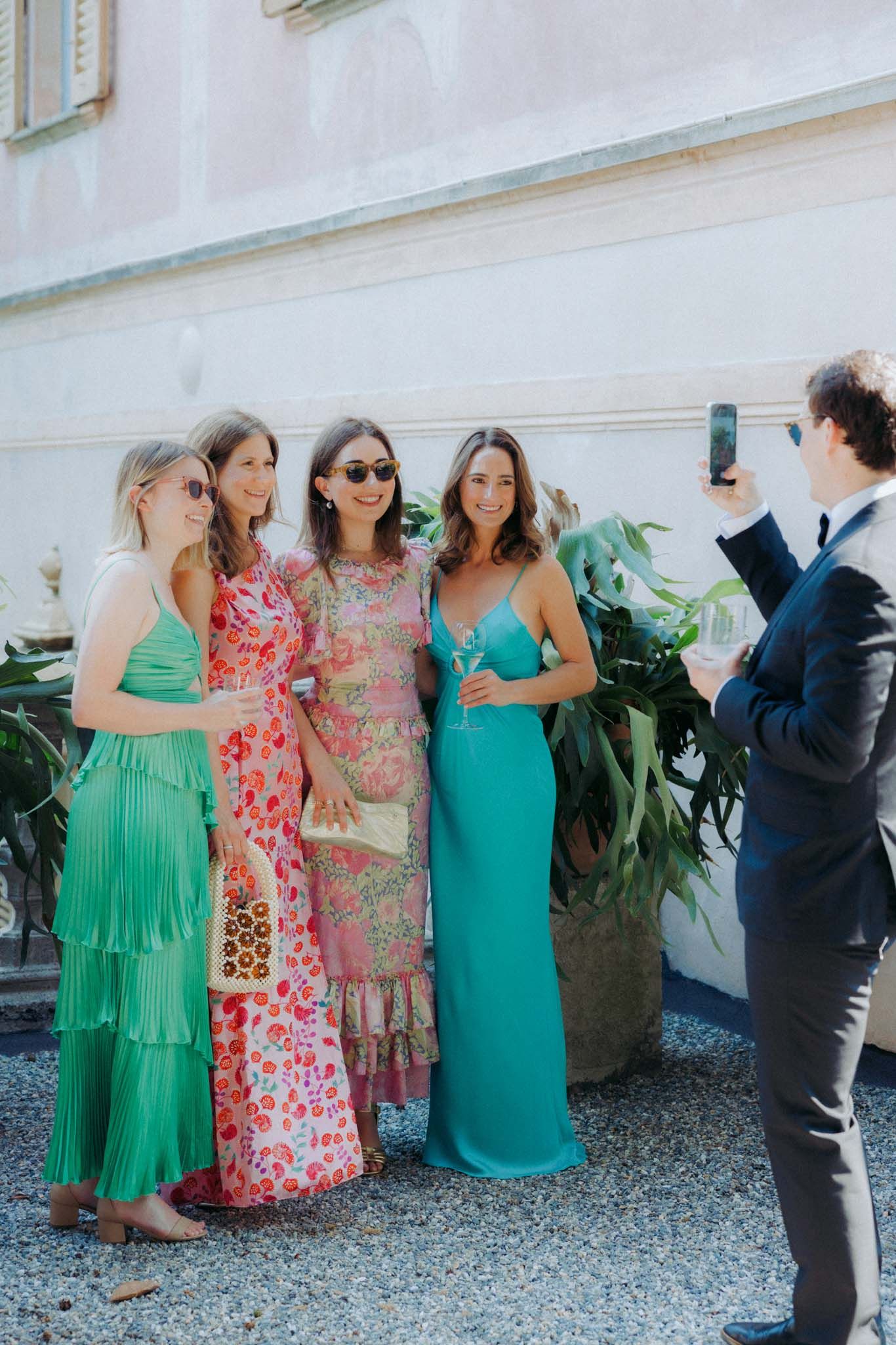 A group of women in green and floral dresses pose together at an outdoor event while someone takes their photo.