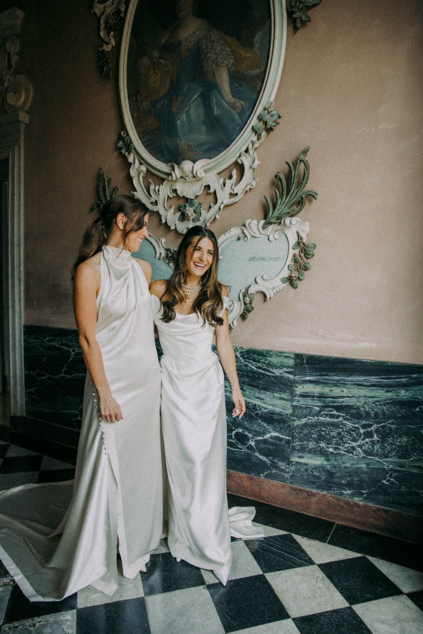 Two people in white dresses share intimate moments on a checkered marble floor against an ornate wall with vintage mirrors.