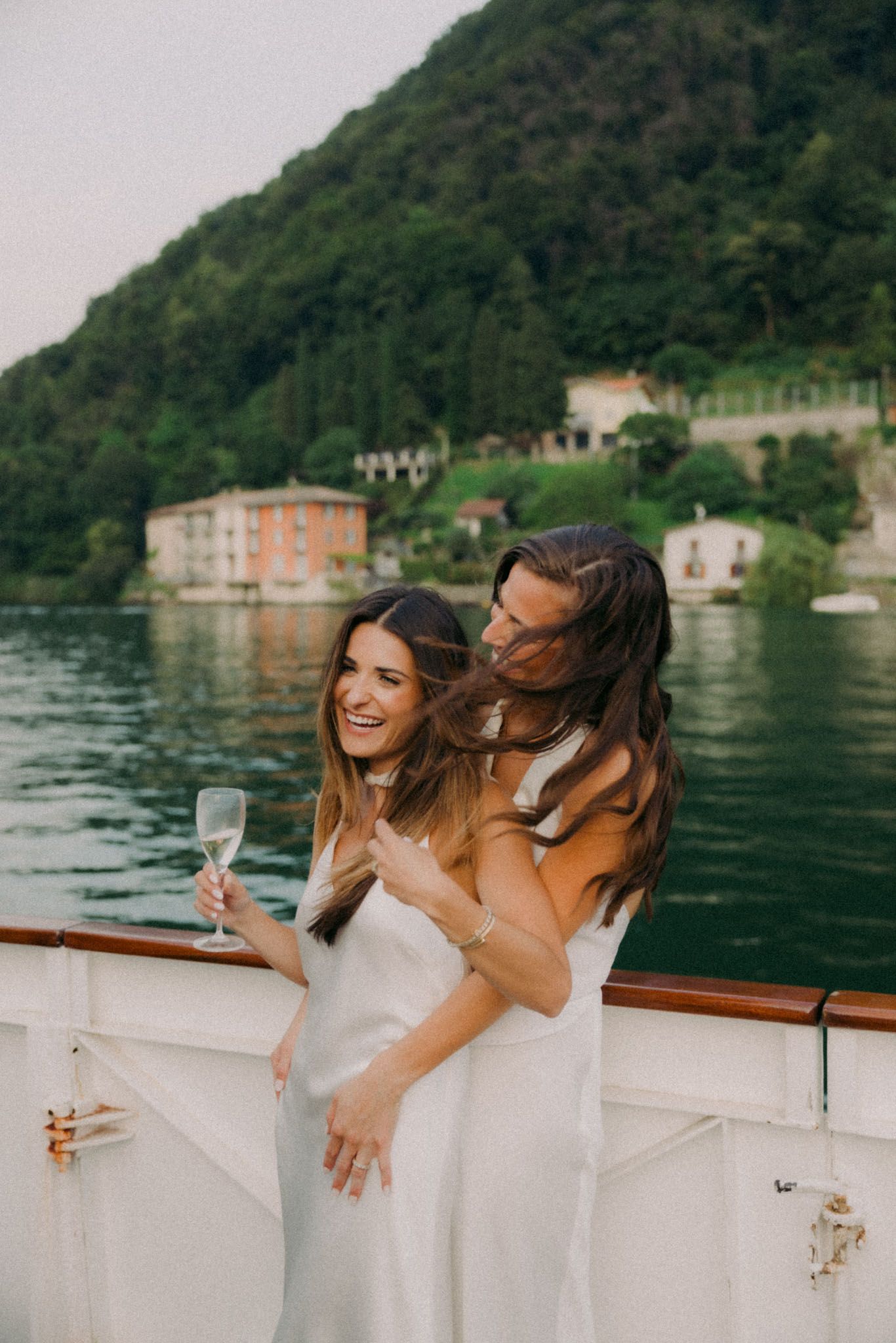 A group of people in white dresses laugh together while enjoying drinks on a boat with mountains in the background.